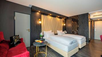 Una habitación de hotel moderna con dos camas, un sillón rojo, una mesita redonda con fruteros y un panel decorativo en la pared.
