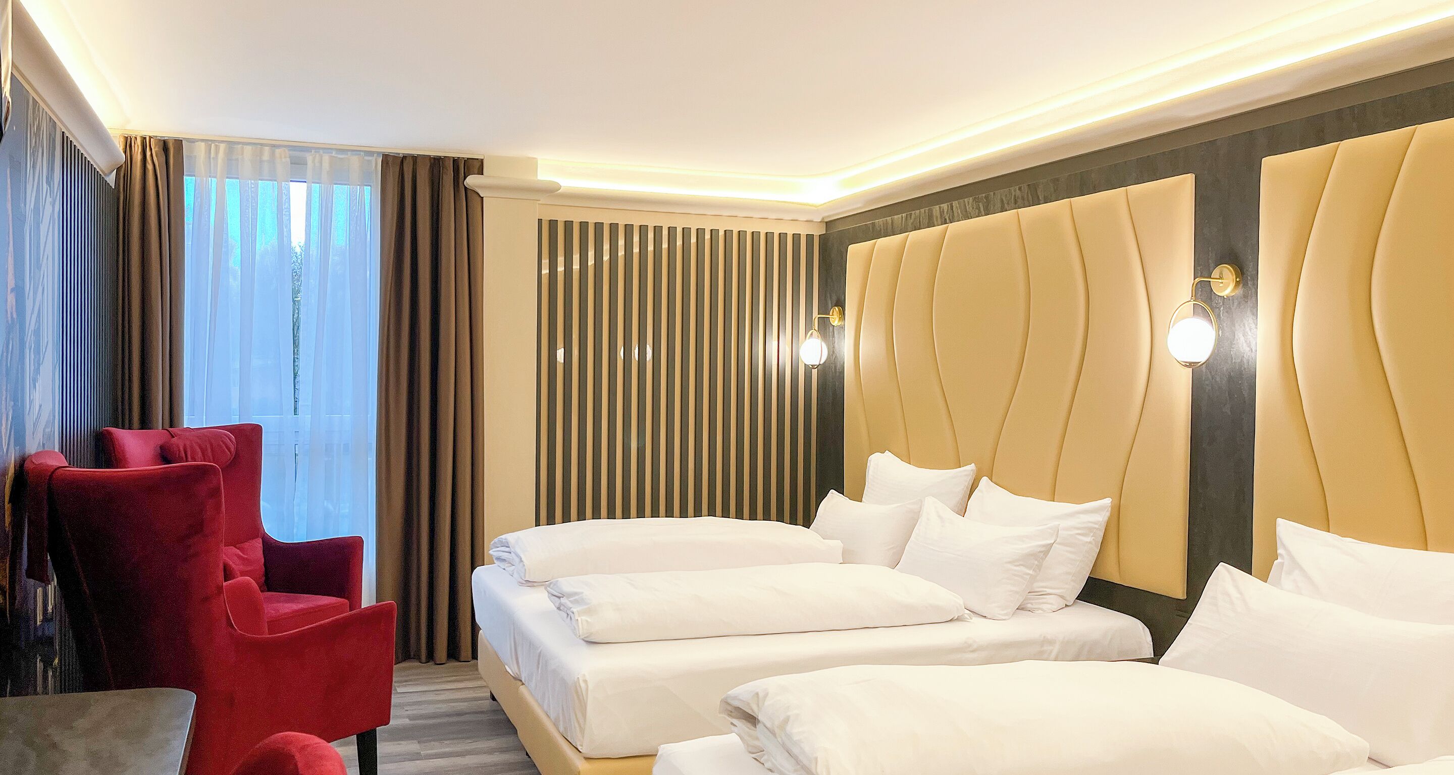 Una habitación de hotel con dos camas, un sillón rojo y un ventanal con cortinas.