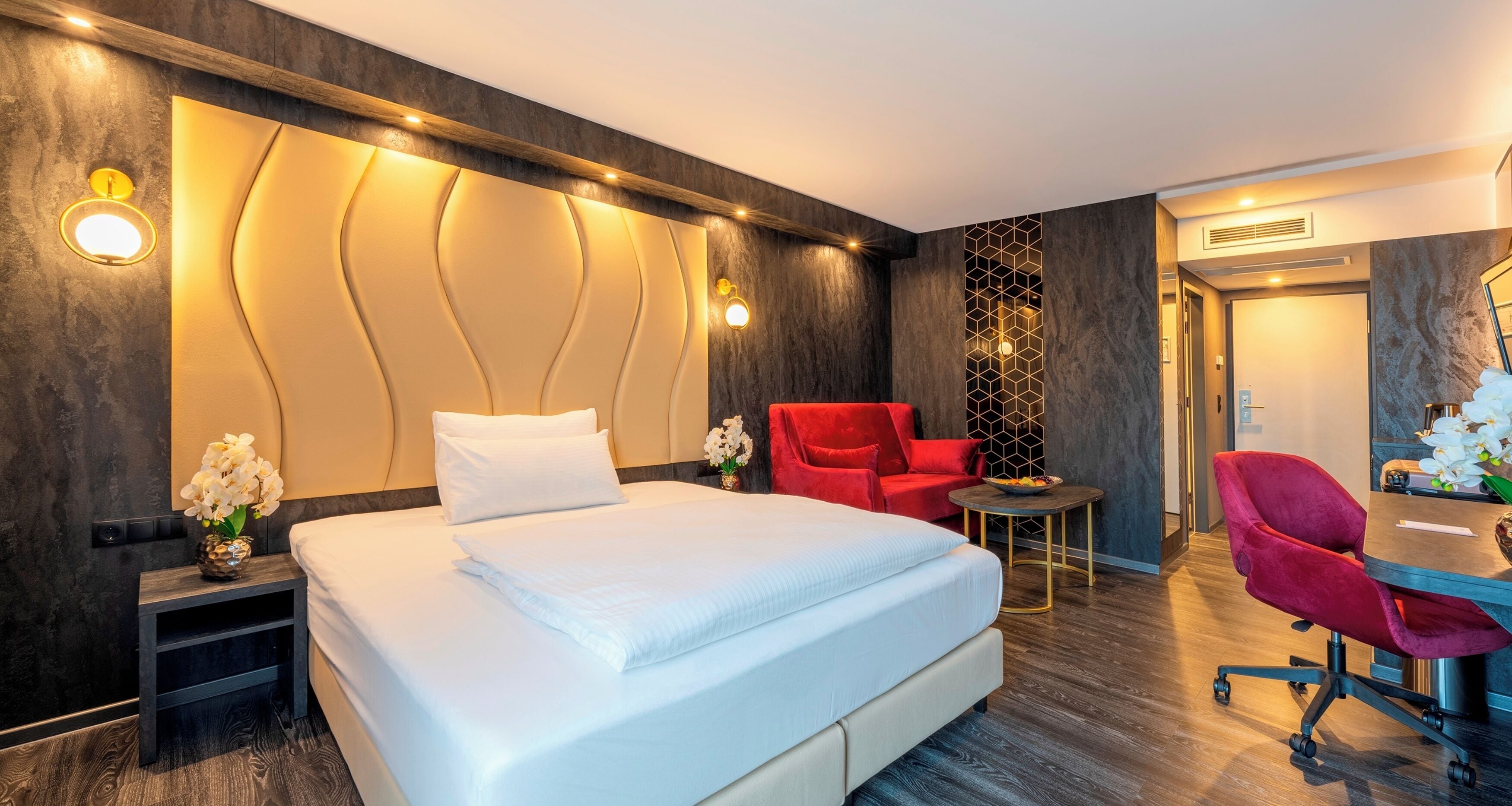 A modern hotel room with a large bed, a desk with a chair, and a red armchair.
