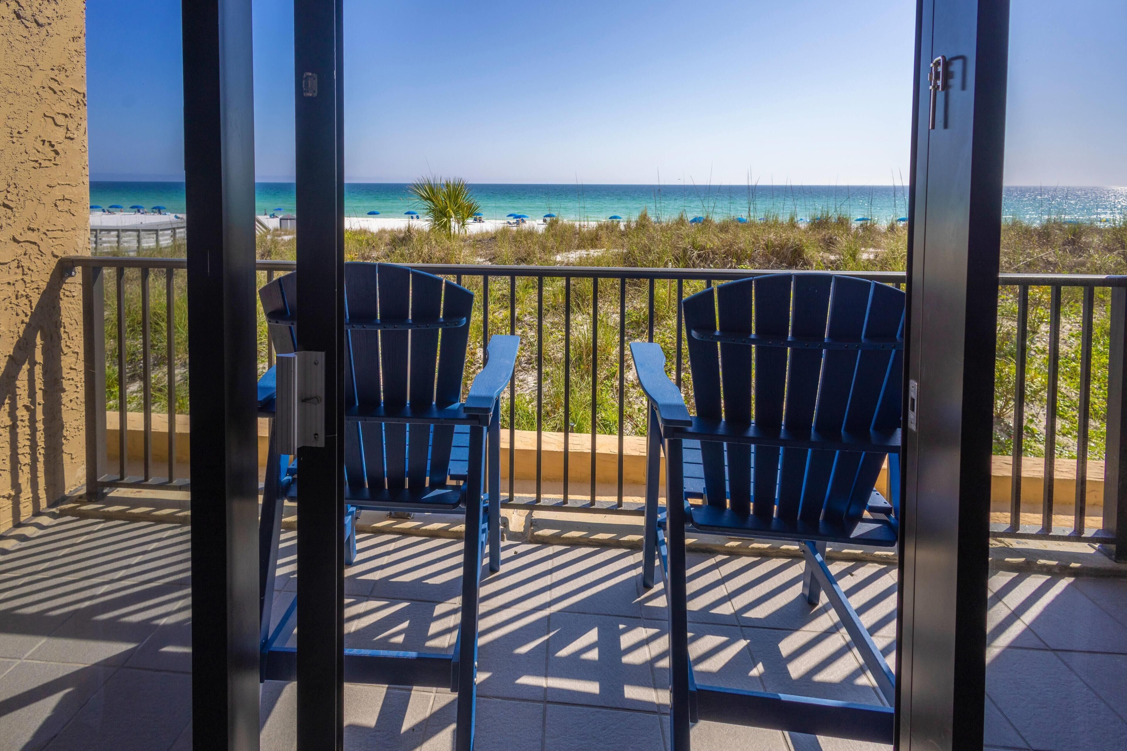 A balcony with two chairs, a view of the beach, and a clear sky.