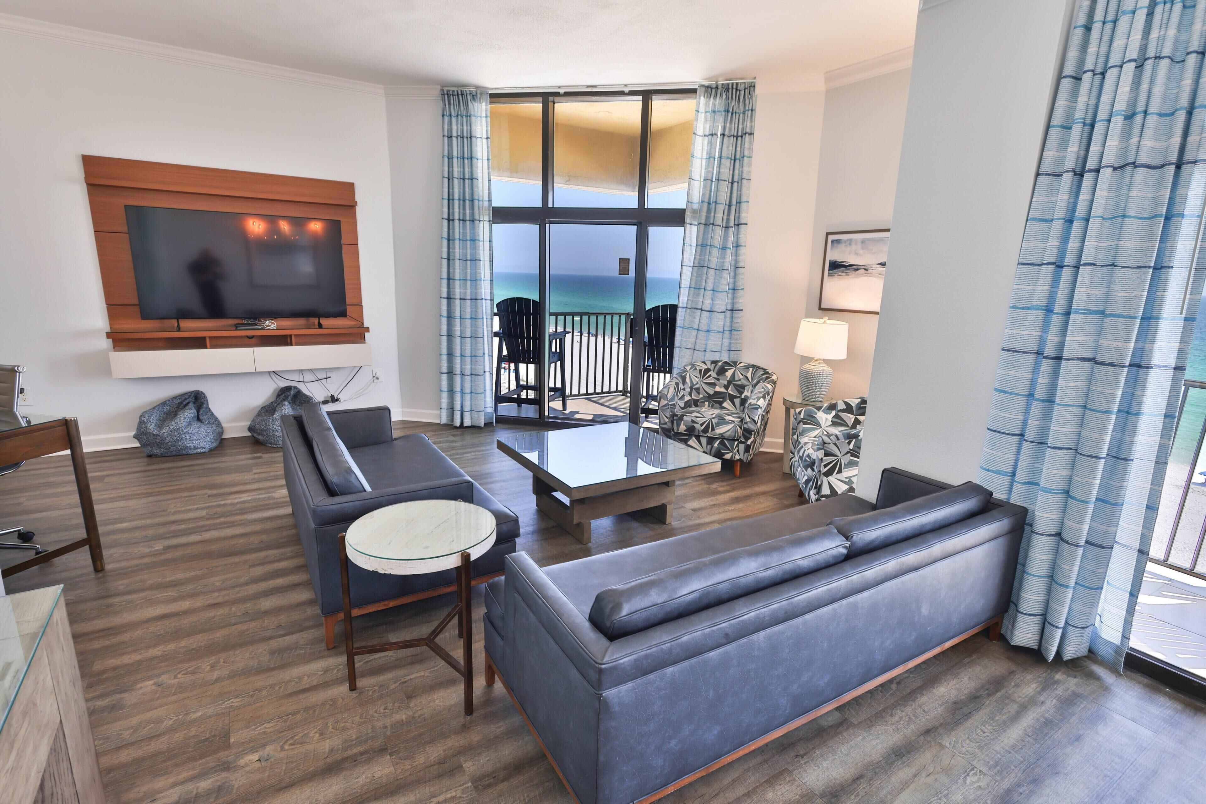A modern living room with a flat-screen TV, a sofa, a glass coffee table, and a view of the ocean through large windows.
