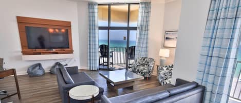 A modern living room with a flat-screen TV, a sofa, a glass coffee table, and a view of the ocean through large windows.
