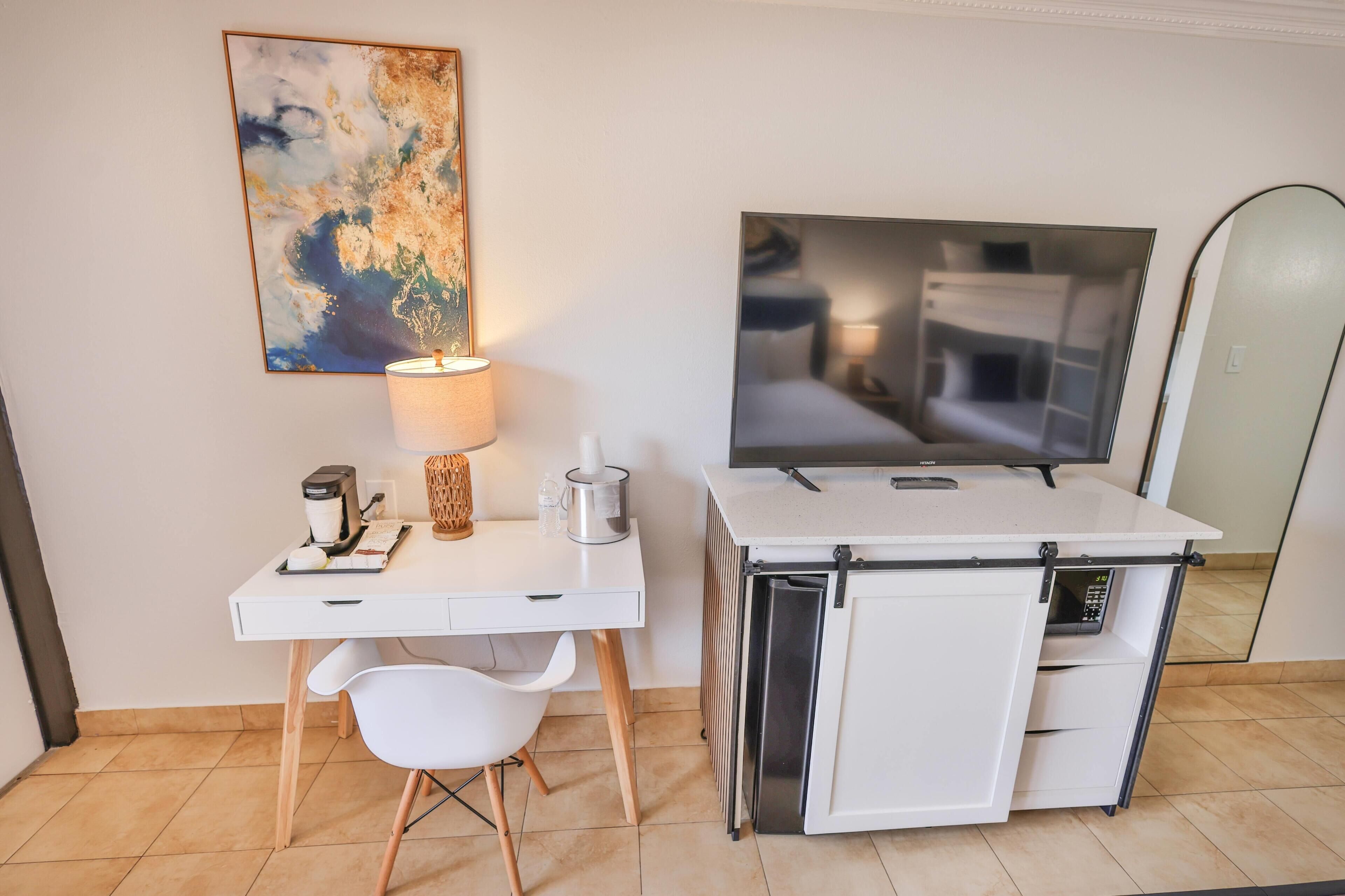 A compact hotel room with a white desk, a chair, a television, a mini-fridge, a coffee maker, and a painting on the wall.
