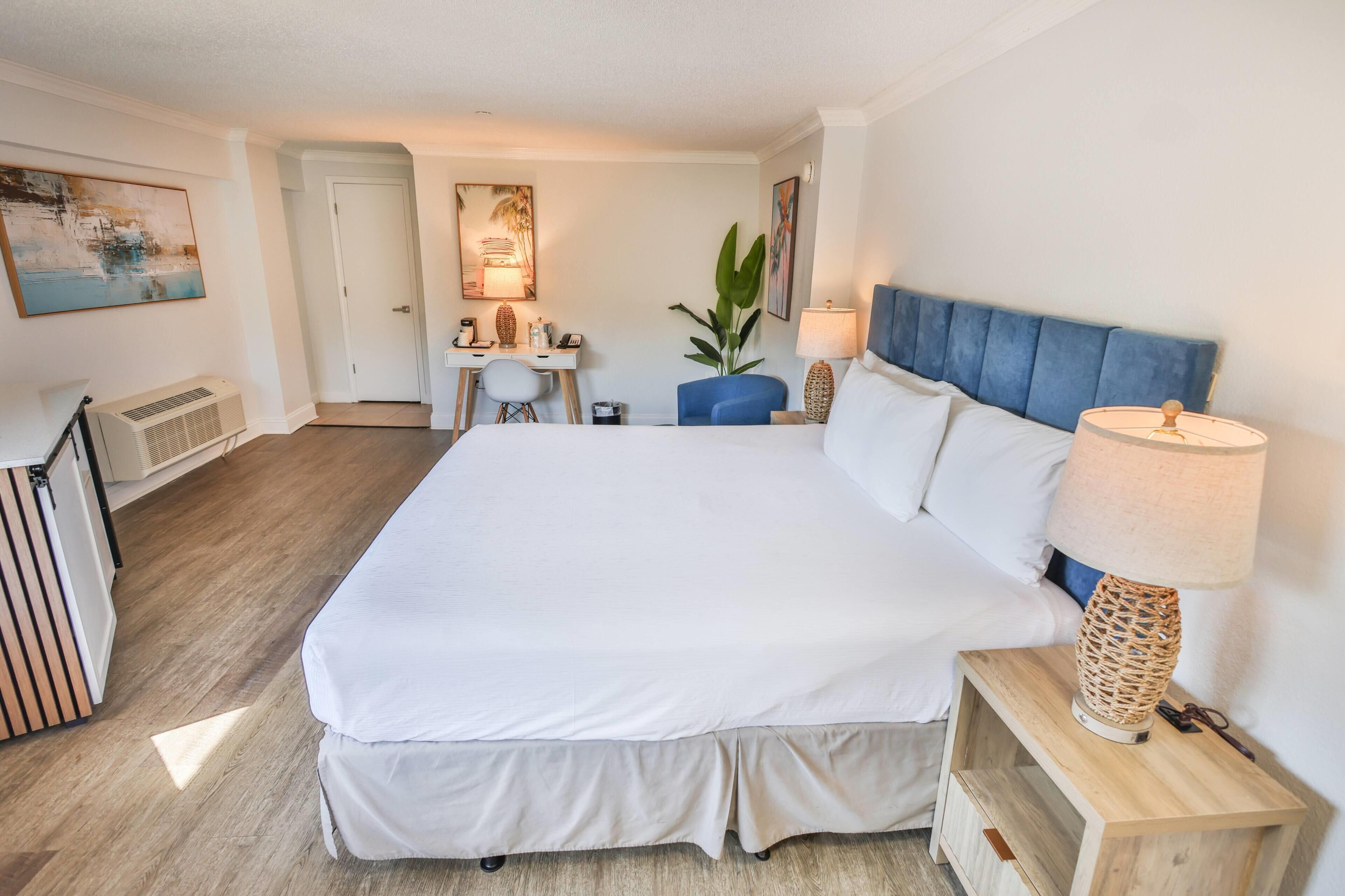 Habitación de hotel con una cama grande, un escritorio, una silla, una planta en maceta y una lámpara.