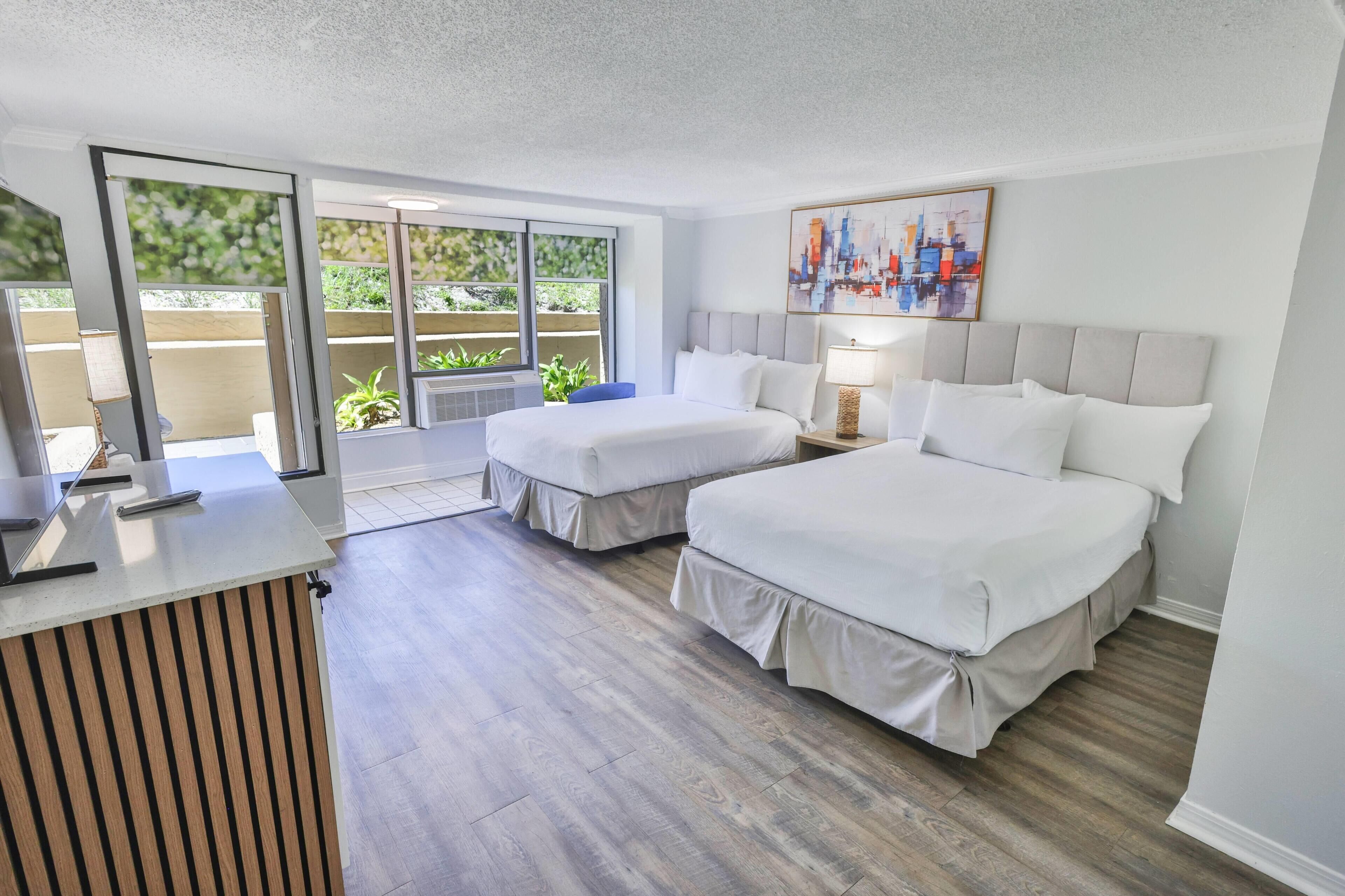 A hotel room with two beds, a desk, a chair, a window with a view, and a painting on the wall.