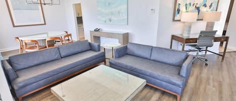 Un salon moderne avec deux canapés gris, une table basse en verre, une table à manger avec des chaises et des œuvres d’art accrochées aux murs.