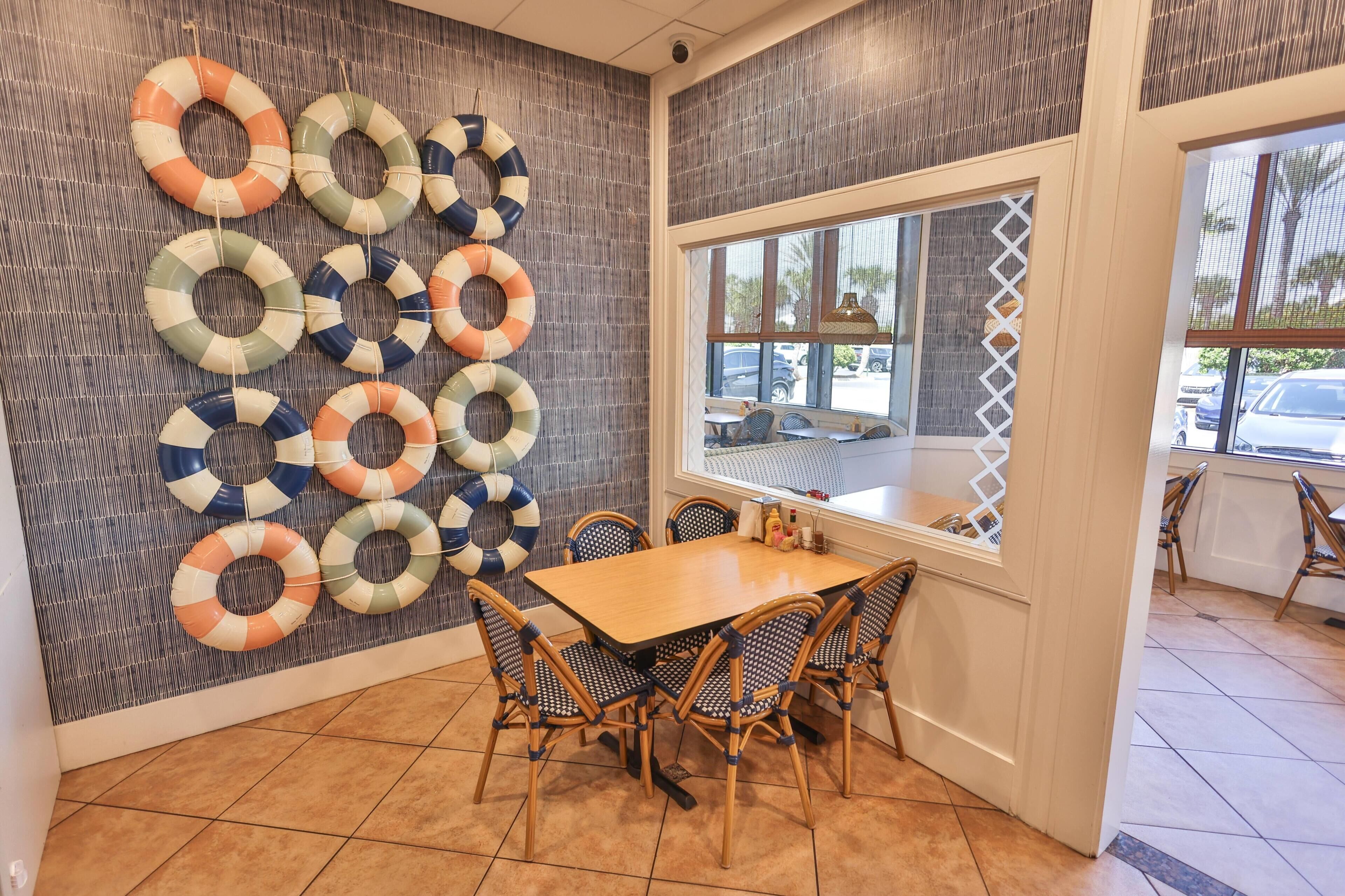 A dining area with a wooden table and wicker chairs, a wall with life rings, and a window with a view of parked cars.