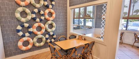 A dining area with a wooden table and wicker chairs, a wall with life rings, and a window with a view of parked cars.