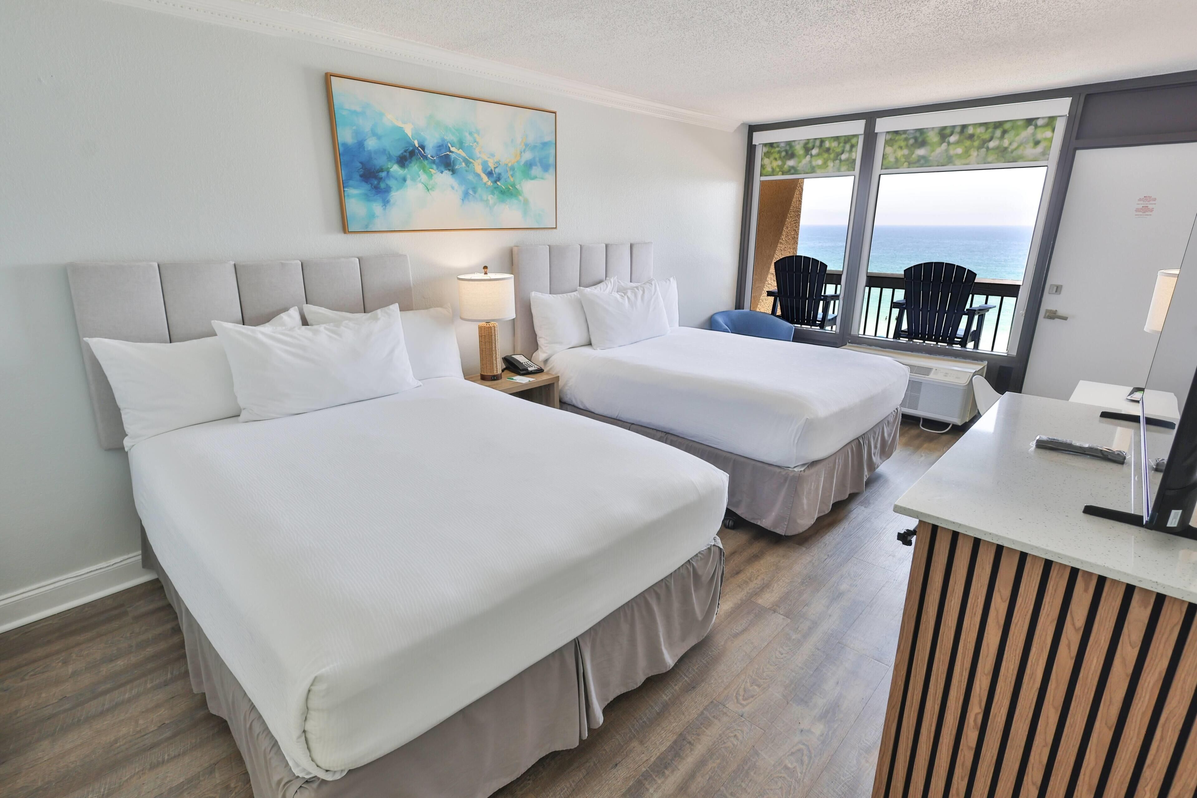 Habitación de hotel con dos camas, un escritorio, una silla y un ventanal con vista al mar.