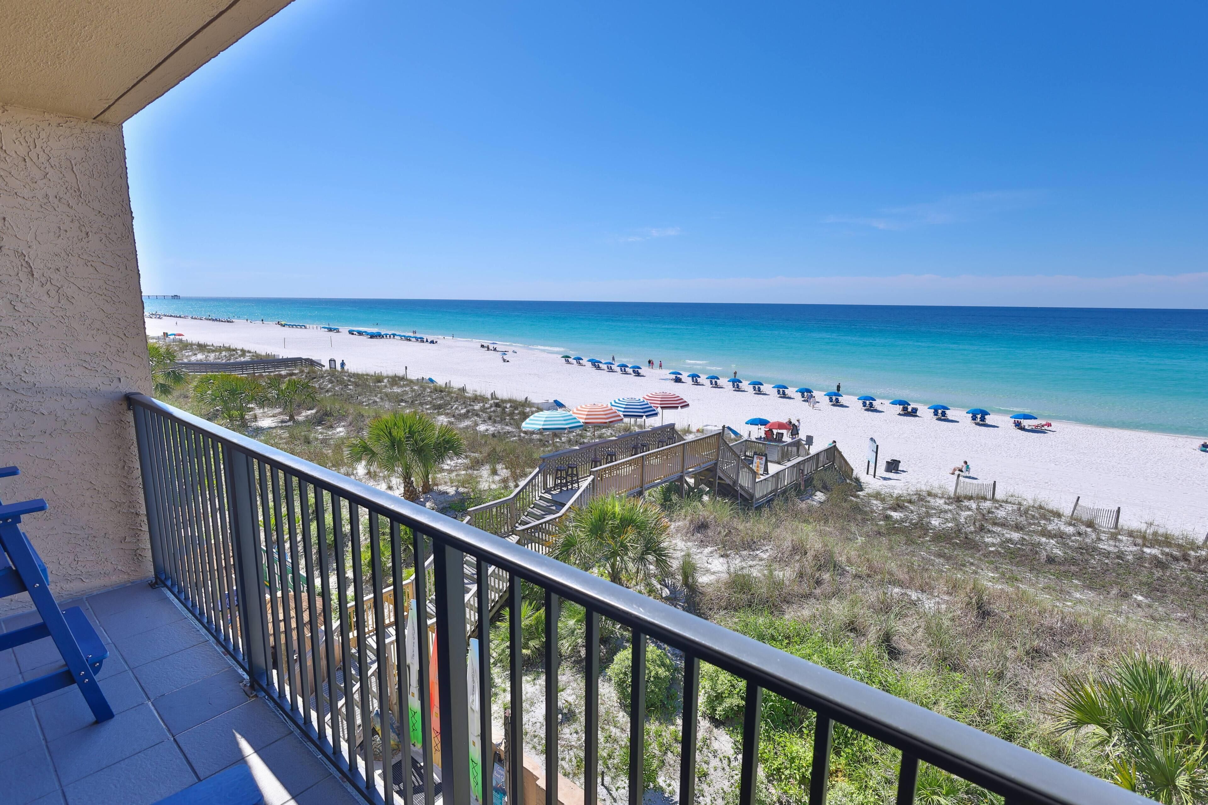 A beach view from a balcony with a wooden deck, beach umbrellas, and a sandy beach.
