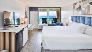 A hotel room with a bed, a desk, a TV, a balcony with chairs, and a large window with a view of the ocean.