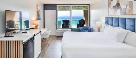 Habitación de hotel con cama, escritorio, televisor, balcón con sillas y un ventanal con vista al océano.