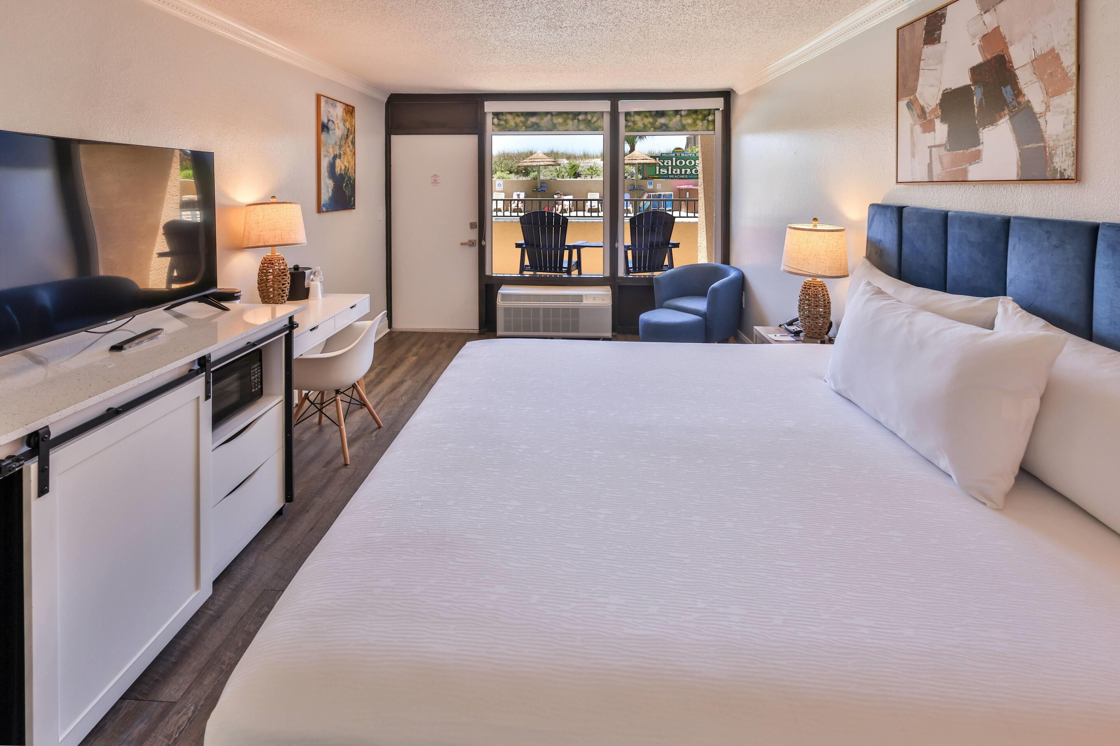 Een hotelkamer met een bed, bureau, televisie en uitzicht op het strand door het raam.