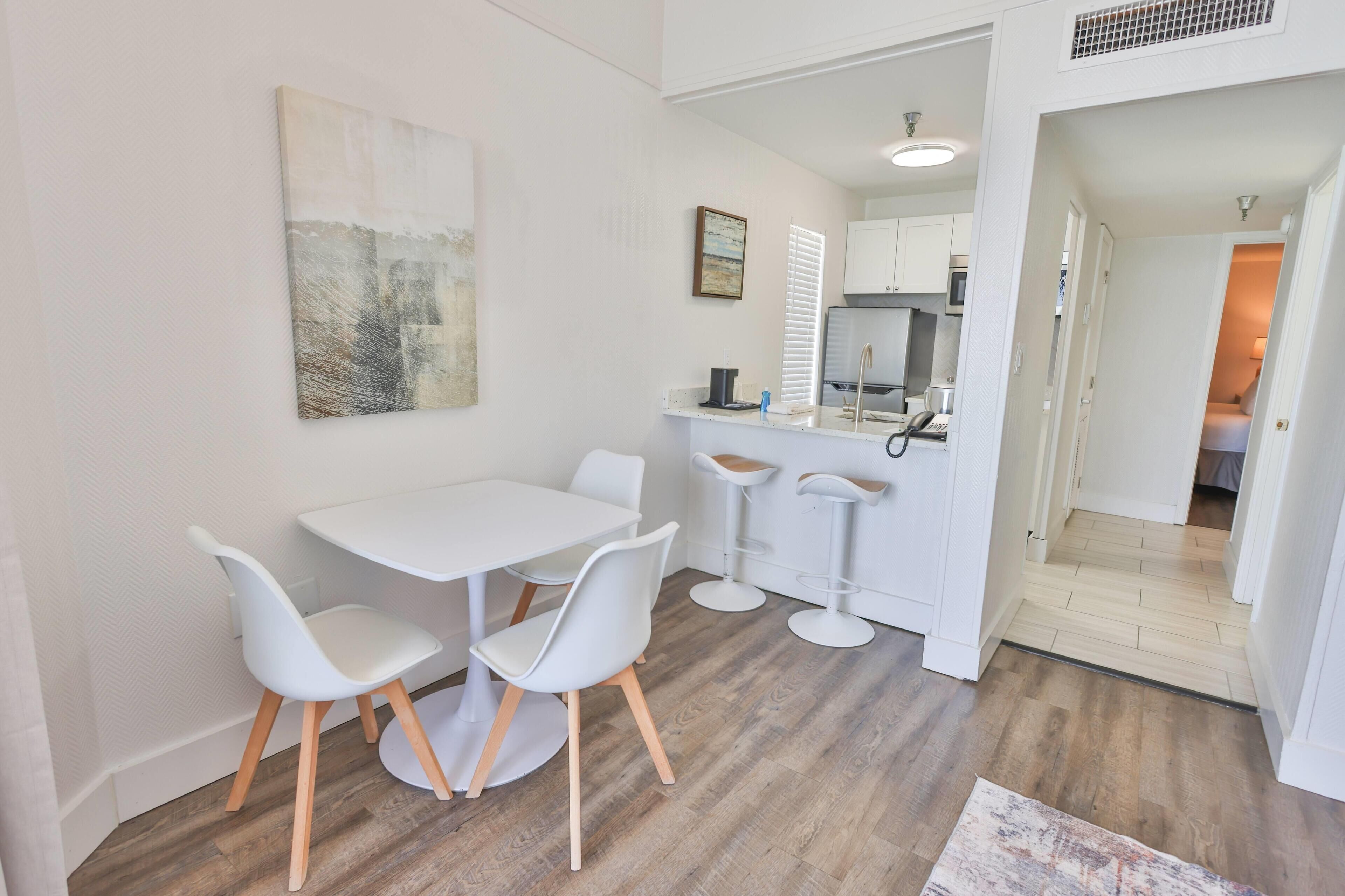 A modern kitchen with white cabinets, a dining area with a white table and chairs, and a hallway leading to another room.