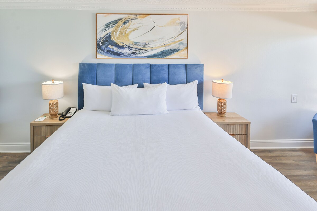 Una cama con cabecera azul, ropa de cama blanca, dos lámparas de noche y un cuadro abstracto colgado en la pared.