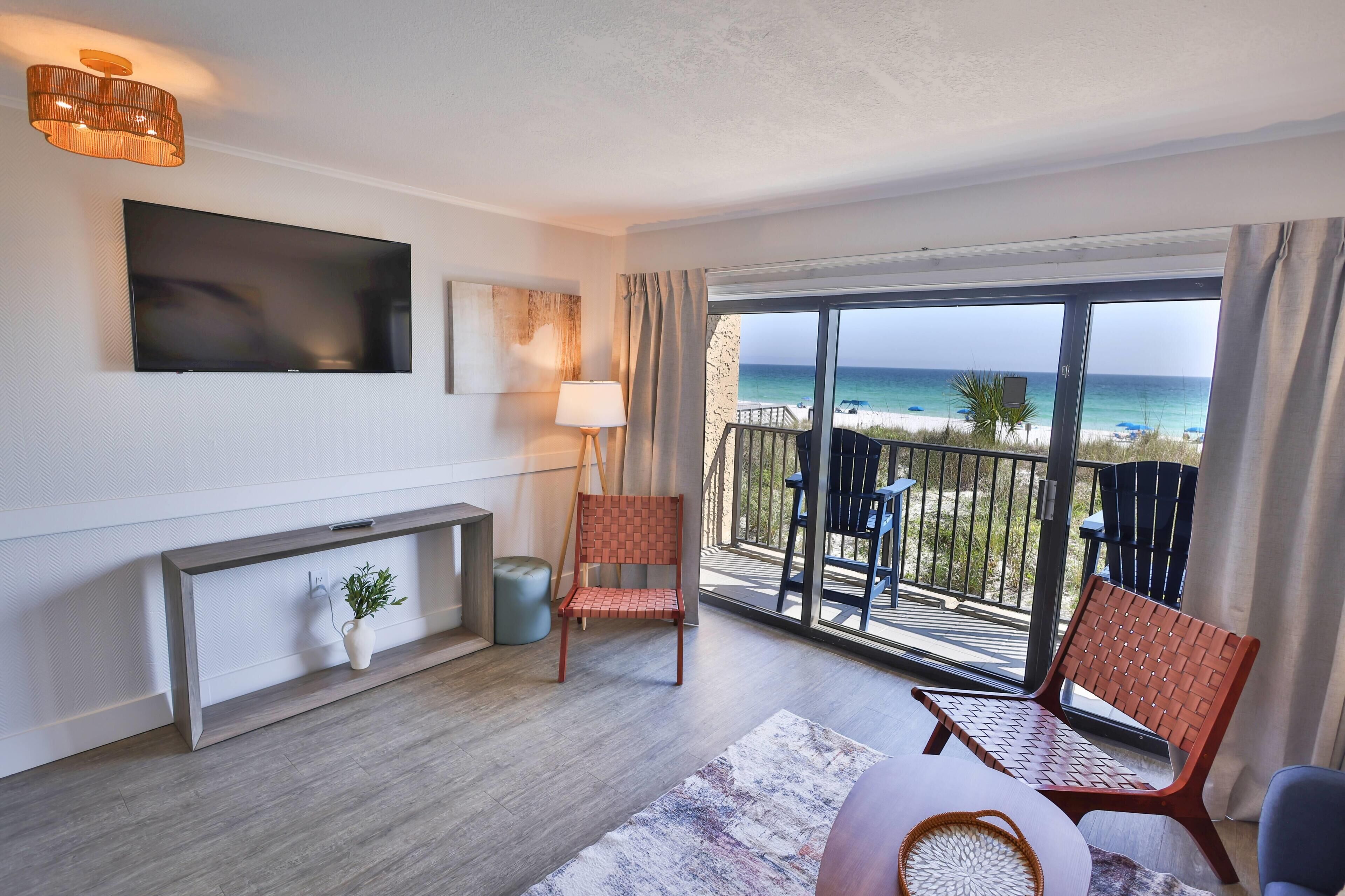 A modern hotel room with a large TV, a fireplace, a balcony with a view of the beach, and two wicker chairs.
