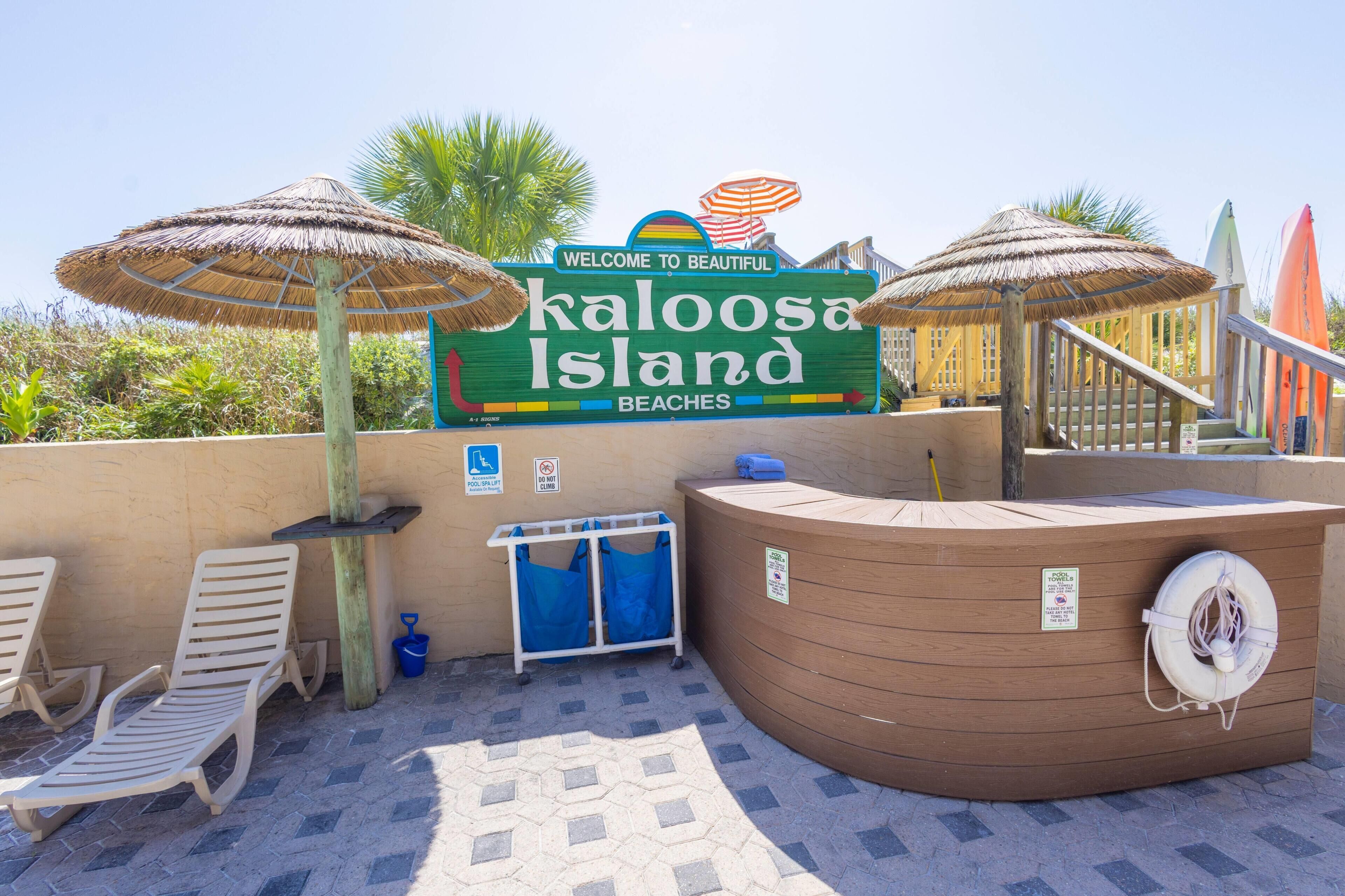 A beachside area with a sign reading 'Welcome to Beautiful Kalooosa Island Beaches,' a hot tub, lounge chairs, and umbrellas.