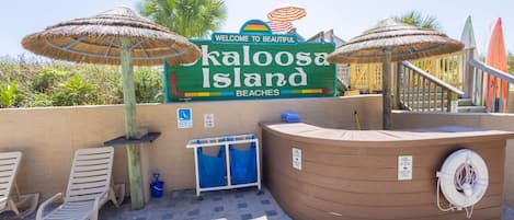 A beachside area with a sign reading 'Welcome to Beautiful Kalooosa Island Beaches,' a hot tub, lounge chairs, and umbrellas.