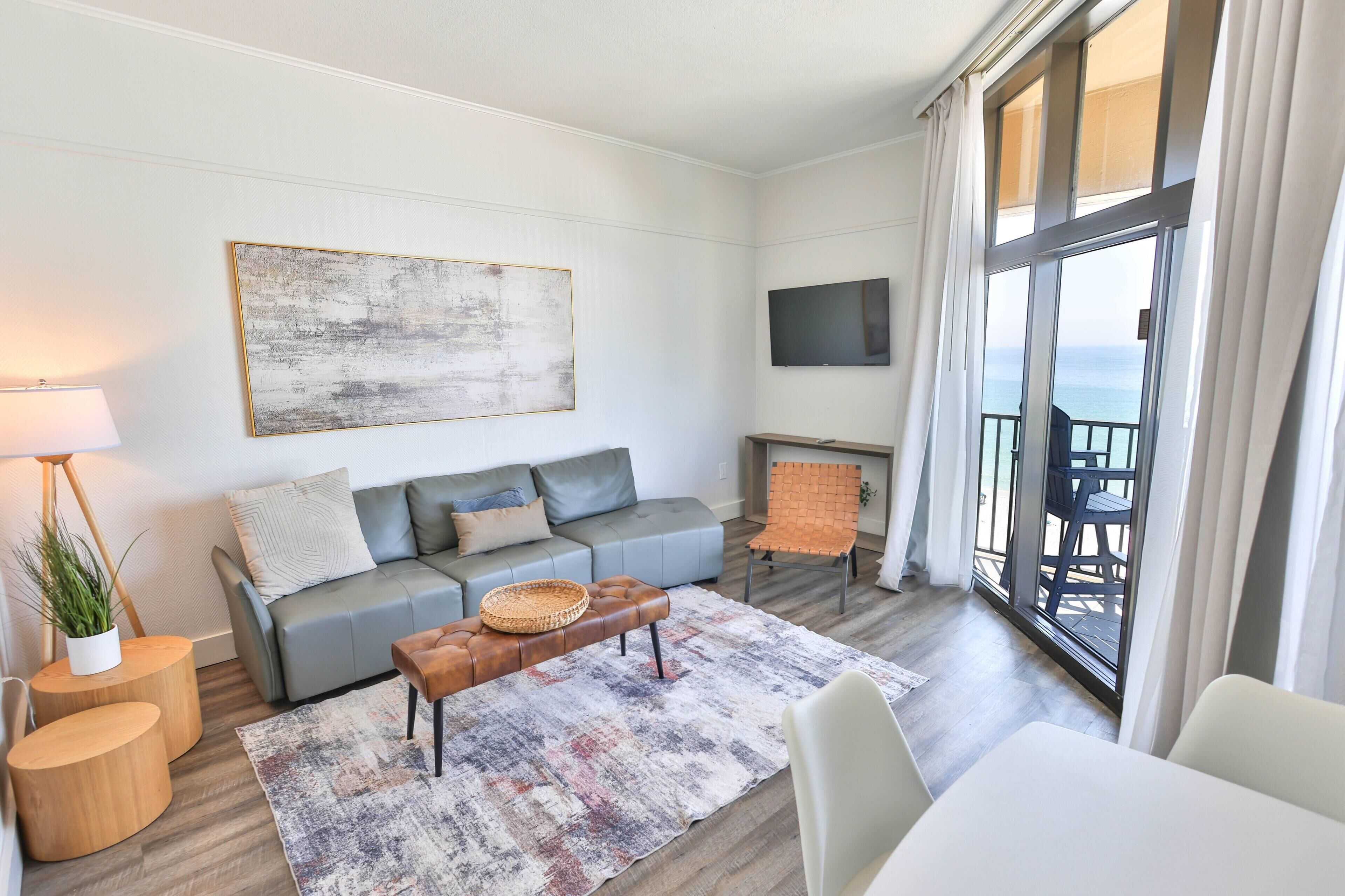 A modern living room with a grey sofa, a coffee table, a flat-screen TV, and a large window with a view.