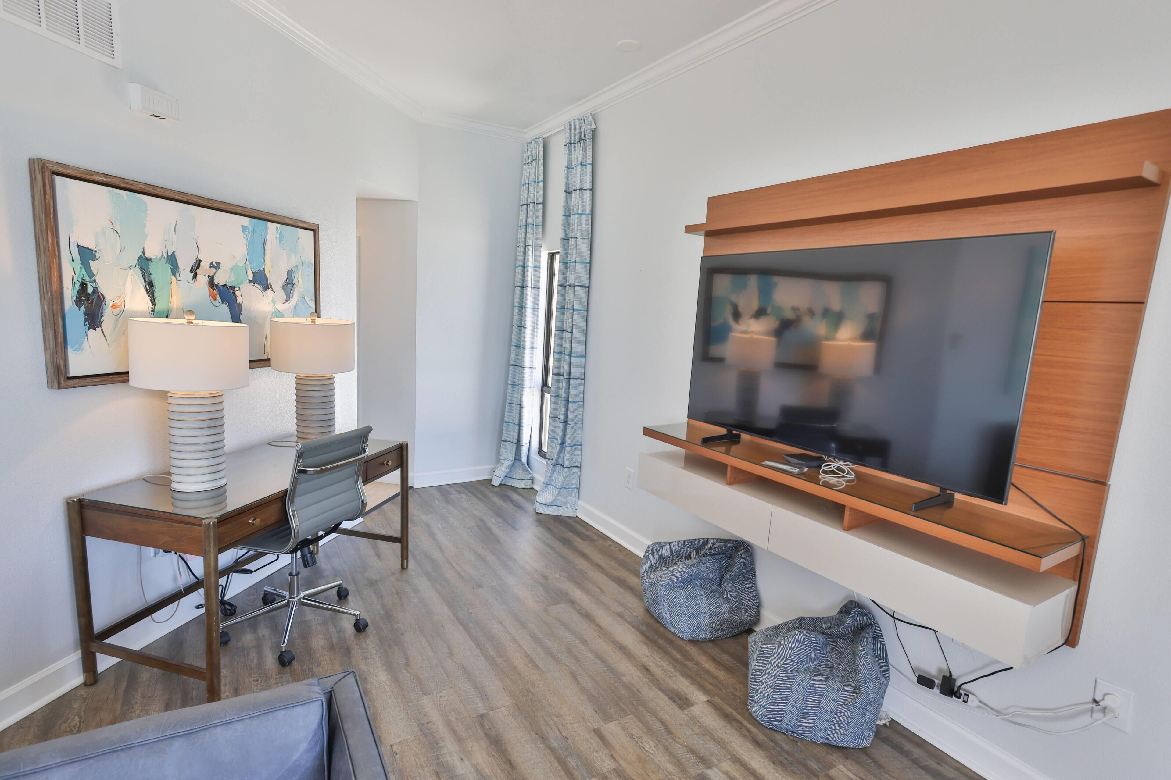 A modern hotel room with a desk, two lamps, a TV mounted on the wall, and a large window with curtains.