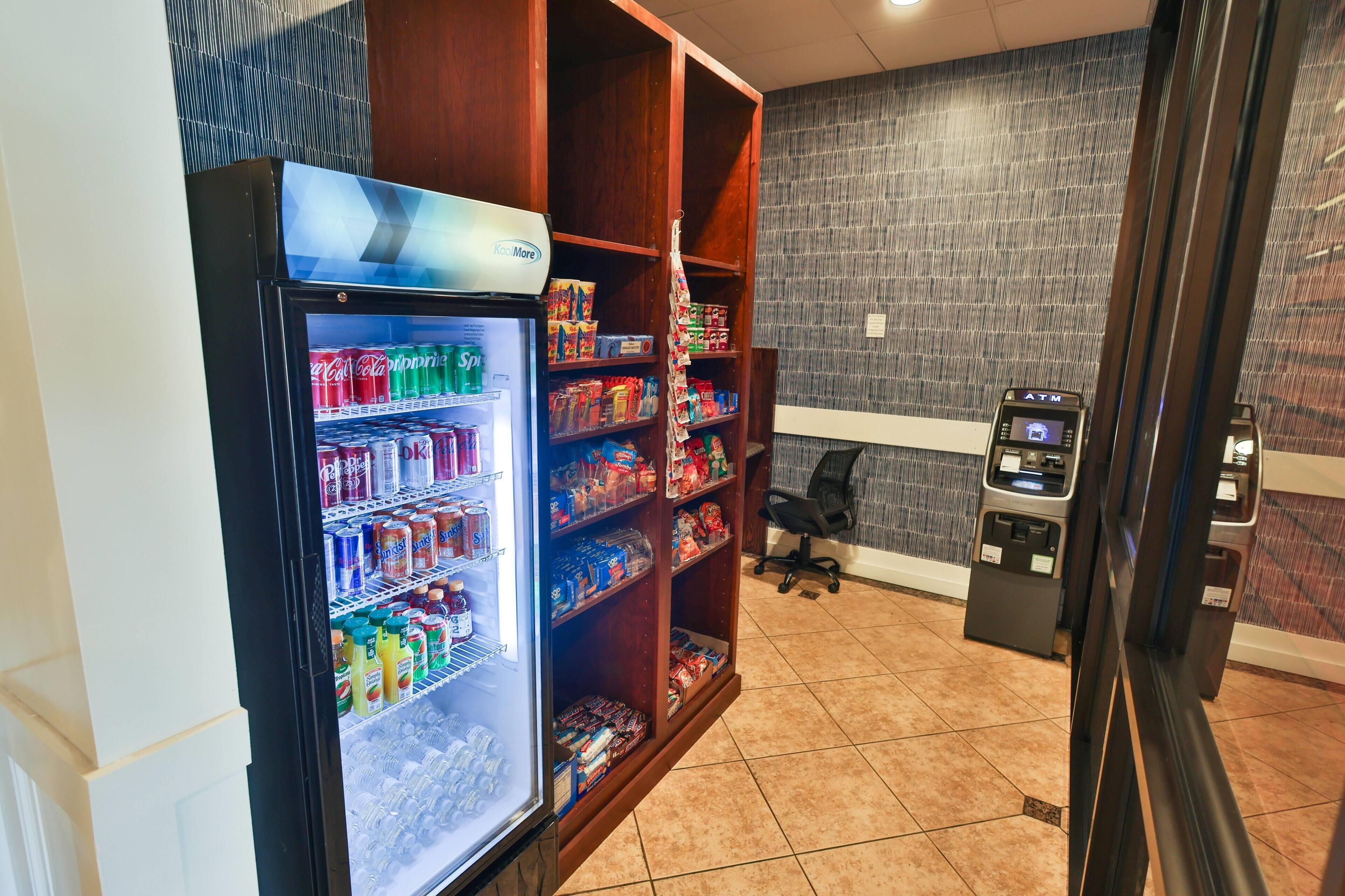 A vending machine, a refrigerator stocked with beverages, and shelves with snacks and other items in a room with tiled flooring and a patterned wall.