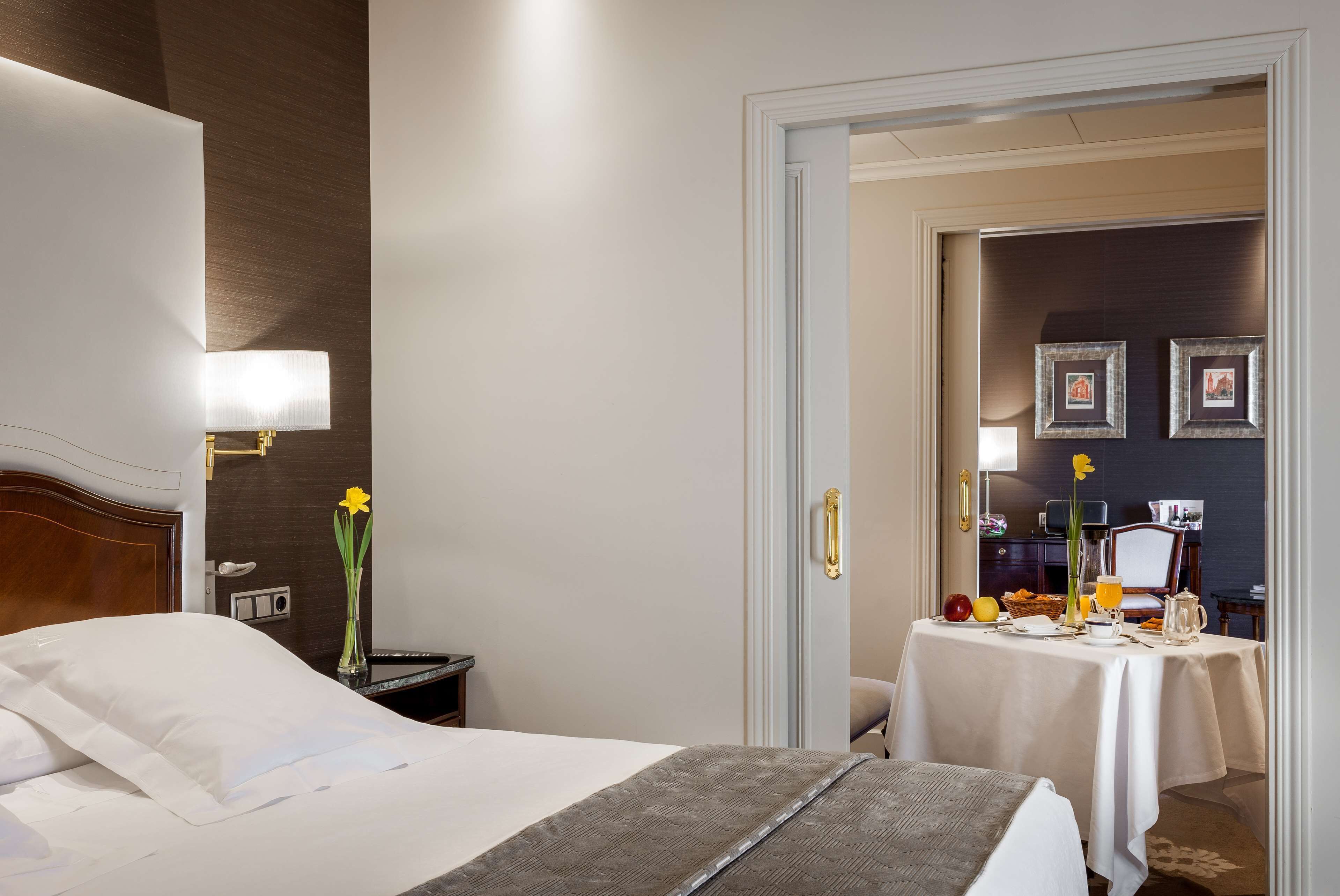 Habitación de hotel con una cama bien hecha, una mesita de noche con lámpara, un jarrón con flores y una mesa de desayuno dispuesta con fruta y una jarra.