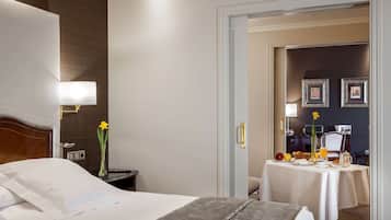 Habitación de hotel con una cama bien hecha, una mesita de noche con lámpara, un jarrón con flores y una mesa de desayuno dispuesta con fruta y una jarra.
