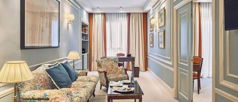 A hotel room with a sofa set, a dining table with a plate of food, a lamp, and a bookshelf.