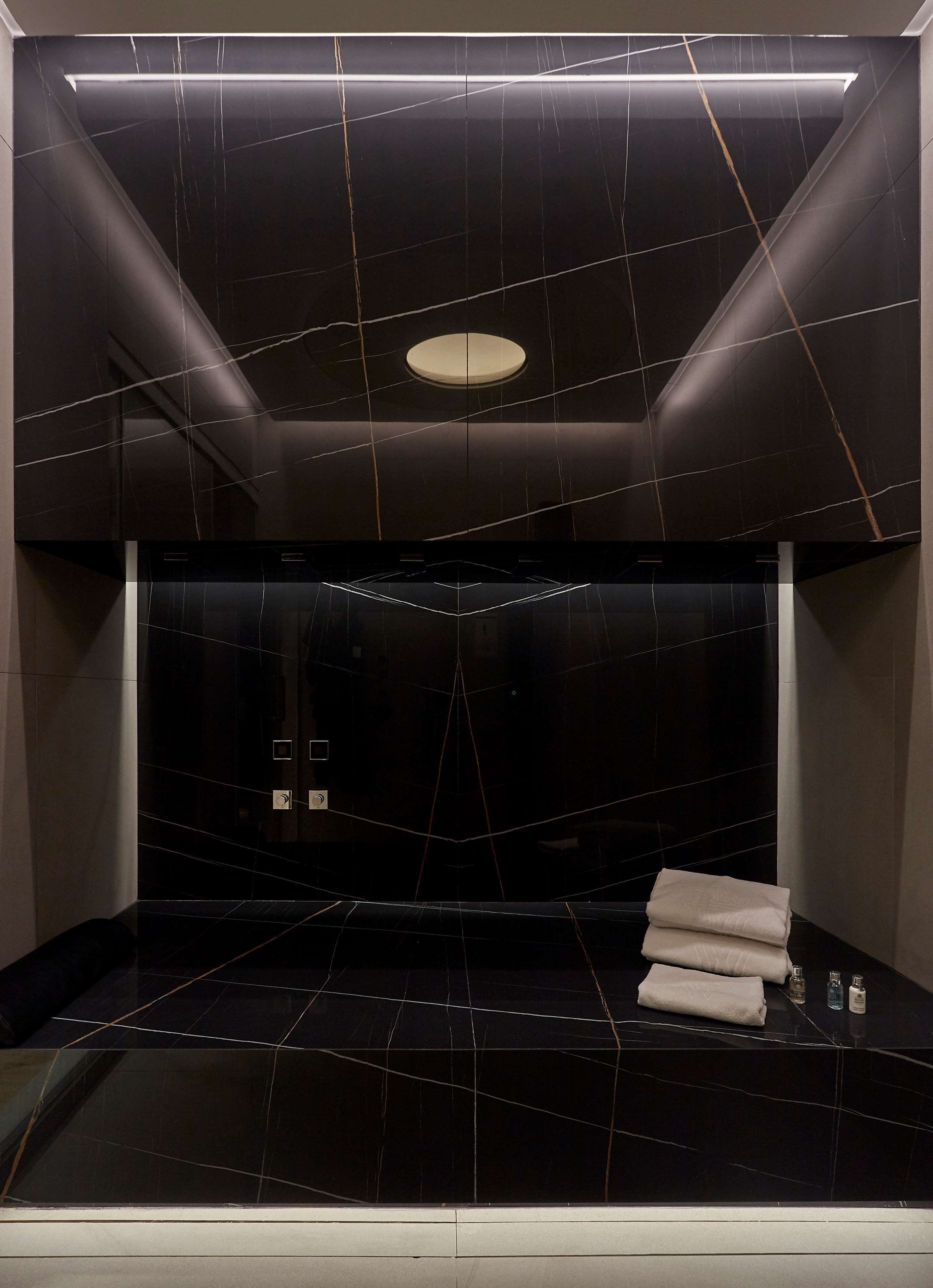 A modern bathroom with black marble tiles, a central light fixture, and a towel rack with folded towels.