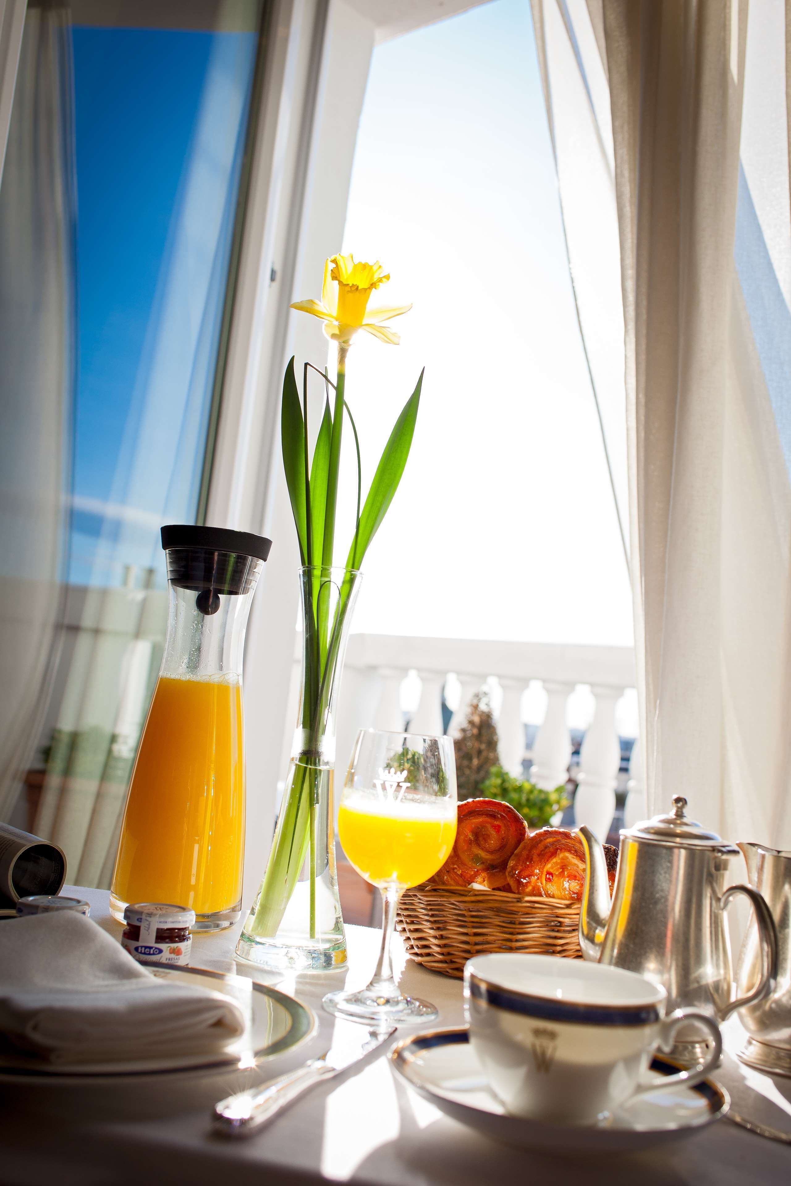 A breakfast table with a glass of orange juice, a glass of orange juice, a cup, a plate, a knife, a spoon, a pitcher, a basket of croissants, and a vase with a flower.