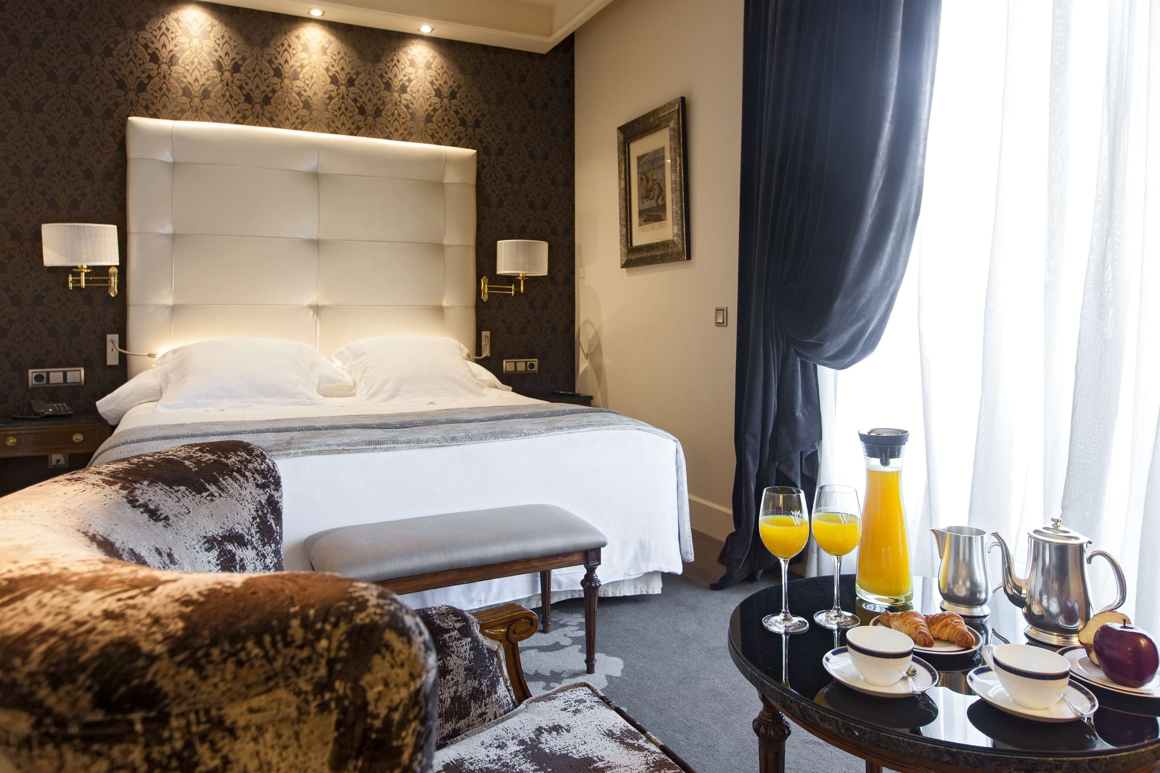 Ein Hotelzimmer mit einem großen Bett, einem Nachttisch mit Lampe, einem Sessel und einem Frühstückstisch mit Croissants, Saft und einer Kaffeemaschine.