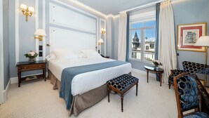 Habitación de hotel con una cama grande, un banco, una mesita con un florero y vistas a un paisaje urbano a través de una ventana.