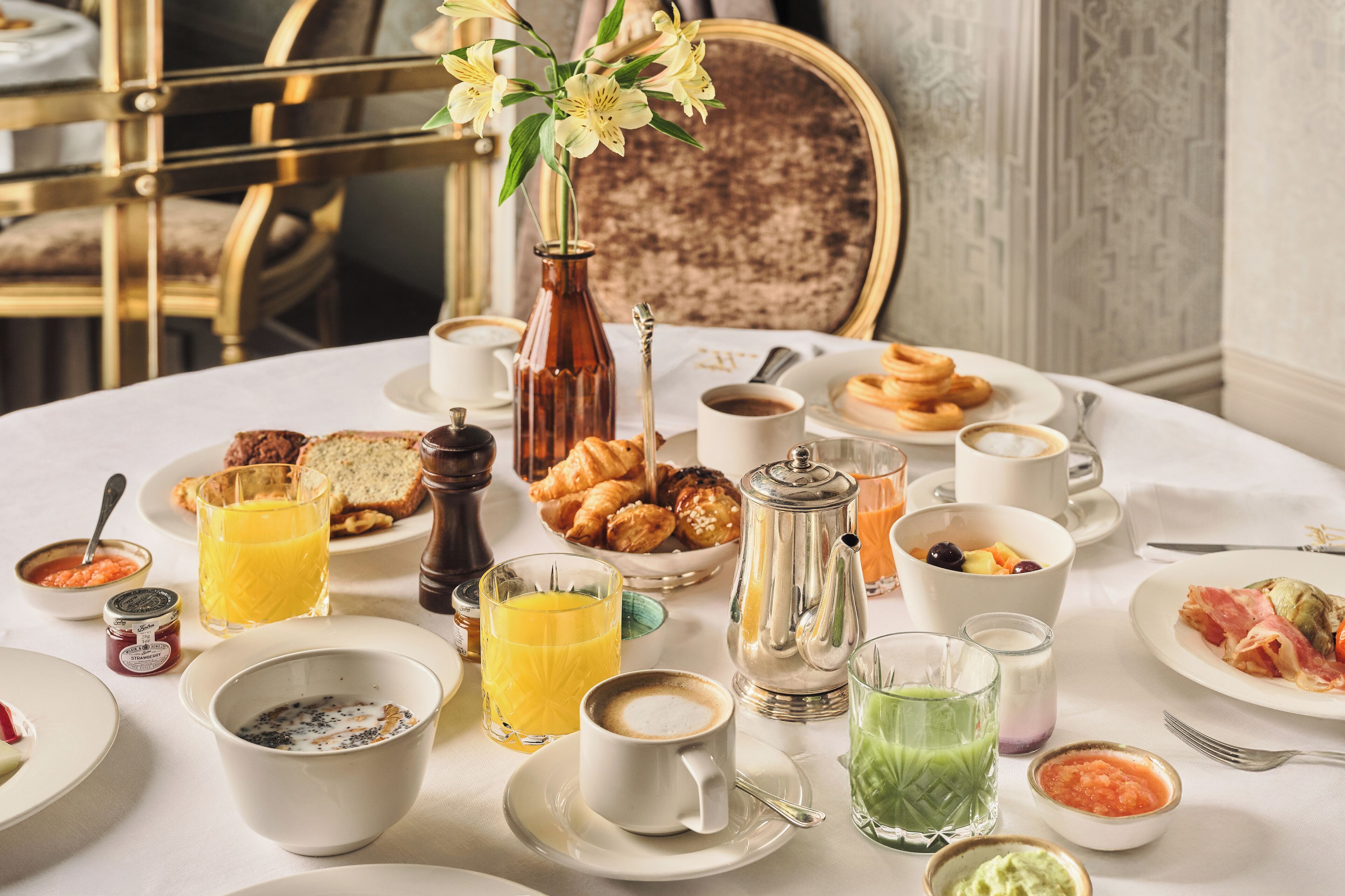 A breakfast table with various dishes, drinks, and a vase of flowers.