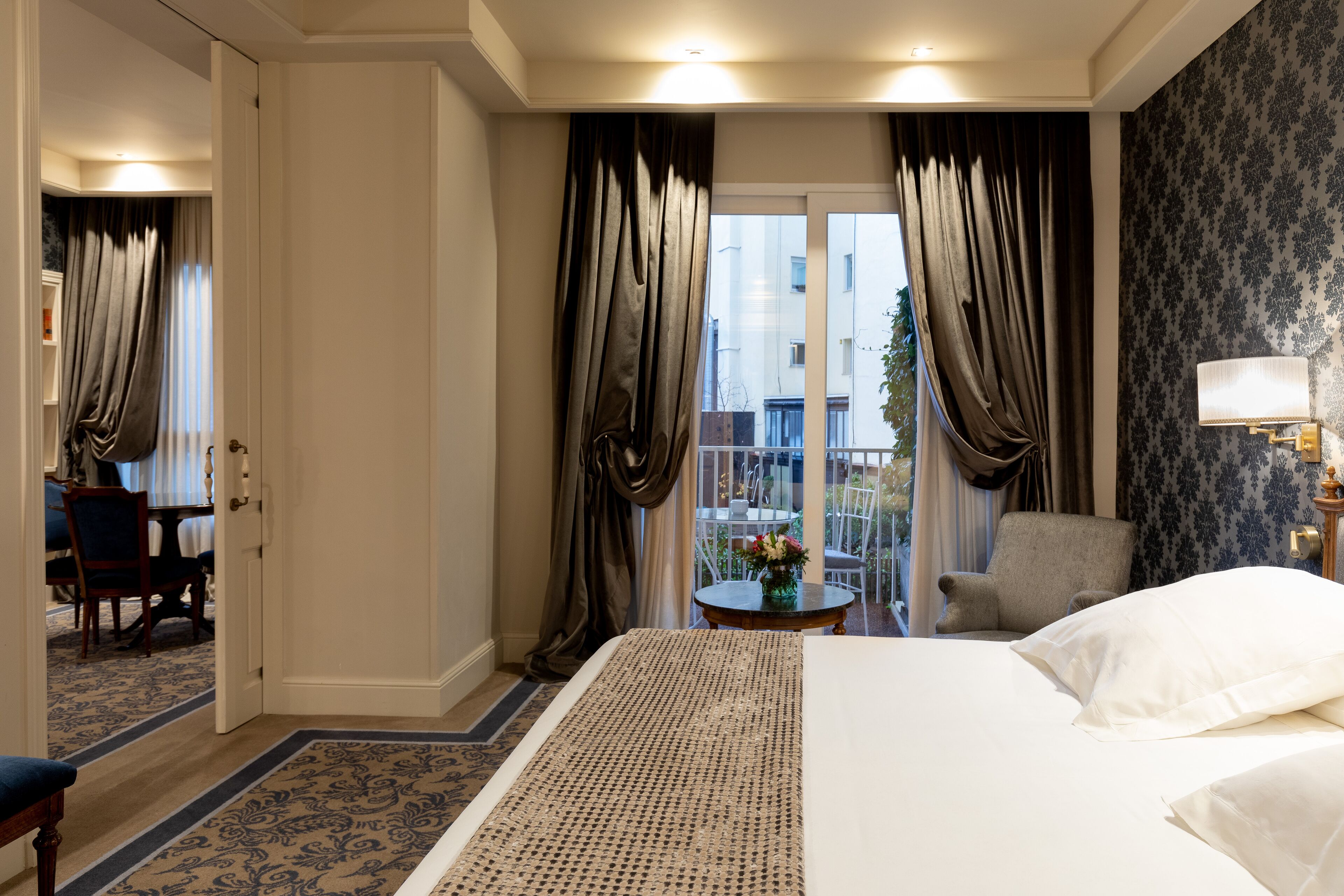 A hotel room with a large bed, a small round table with a vase of flowers, a chair, and a balcony with curtains.