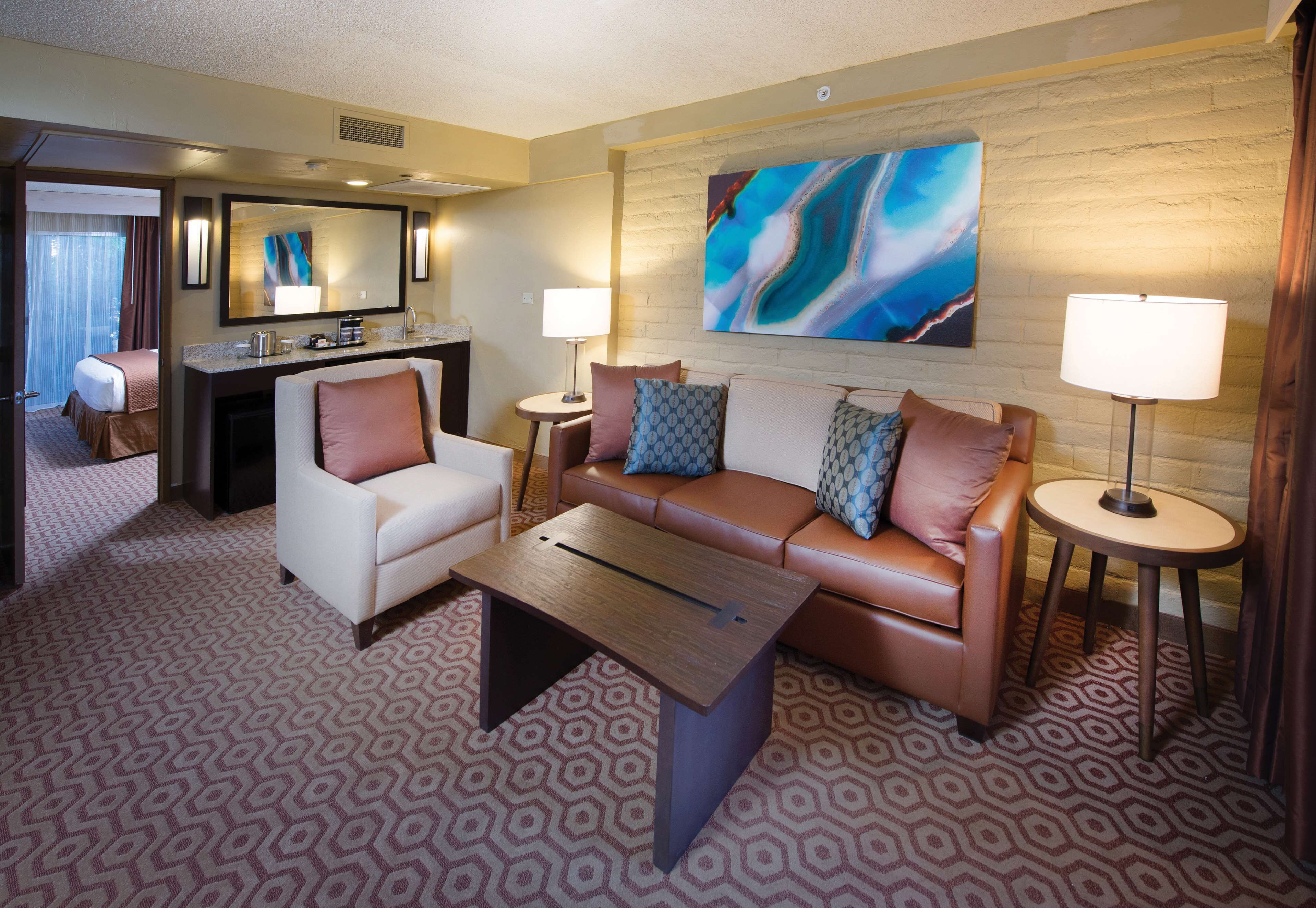 A hotel room with a sofa, armchair, coffee table, and a dining area with a small table and chairs.