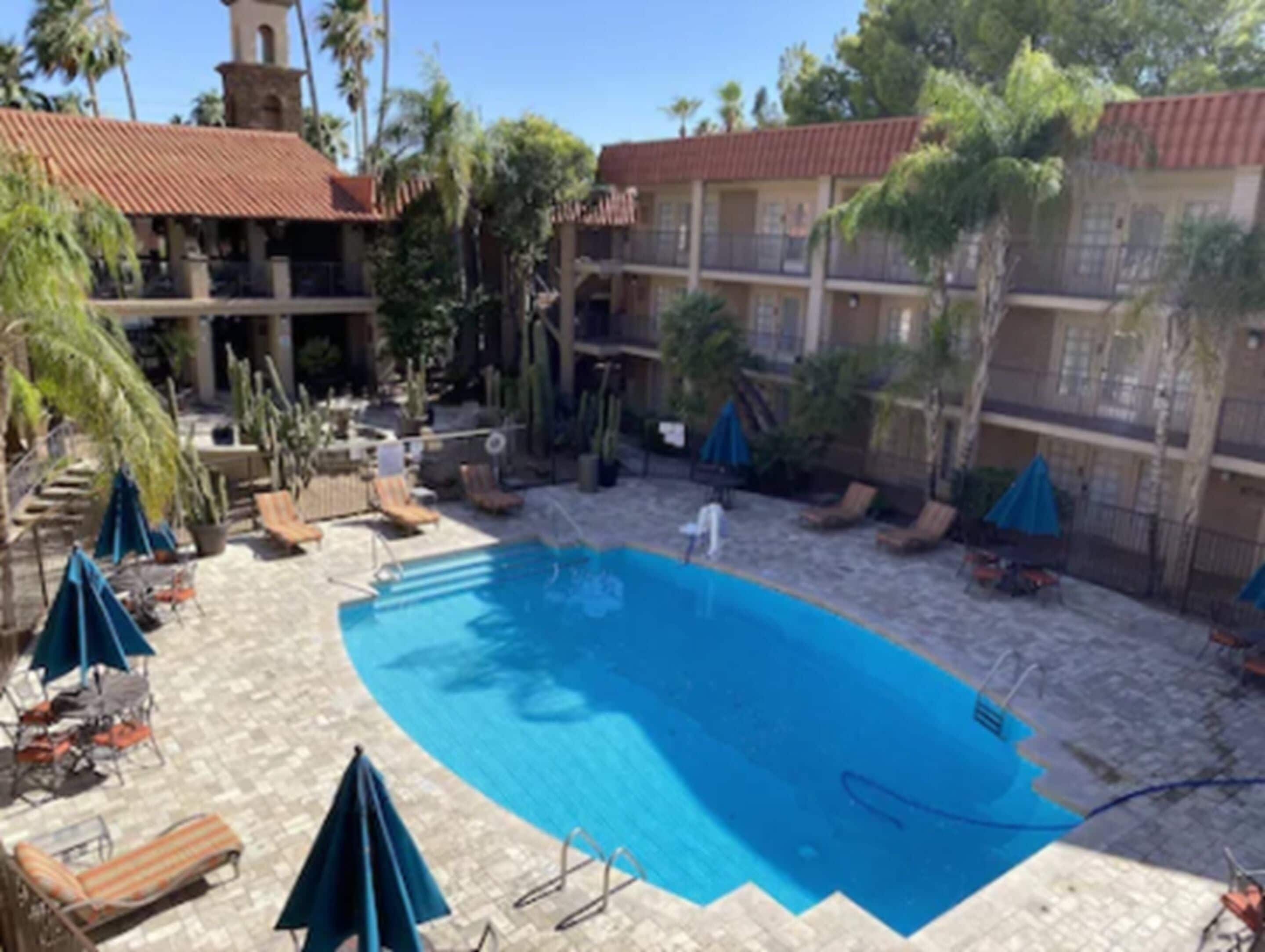A pool area with lounge chairs and umbrellas, surrounded by a building with a tiled roof and palm trees.