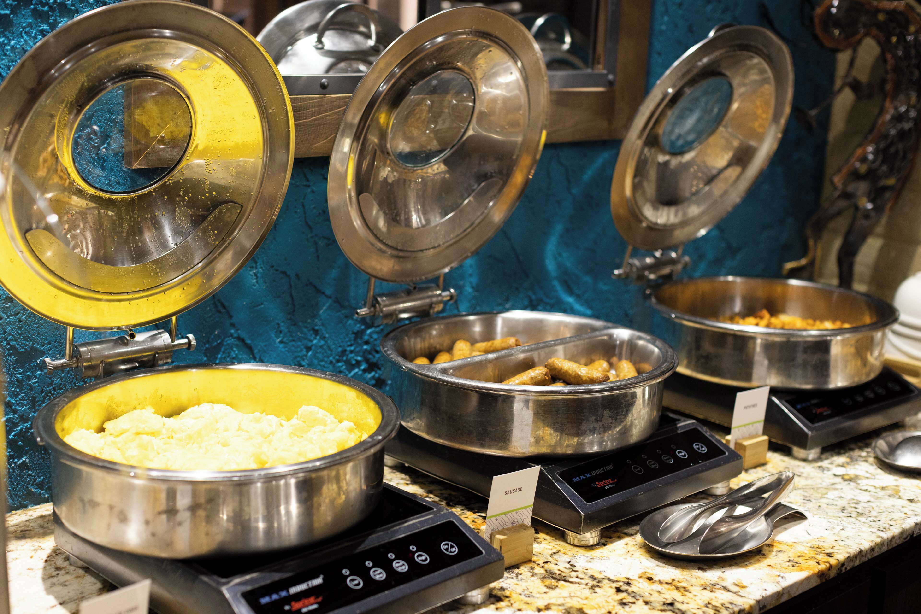 Servicio de comidas tipo buffet con diversos platos en recipientes metálicos, una balanza digital y un cartel que indica 'SAUSAGE'.
