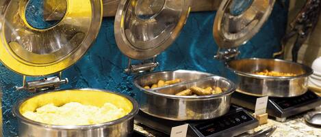 Buffet-style food service with various dishes in metal containers, a digital weighing scale, and a sign indicating 'SAUSAGE'.