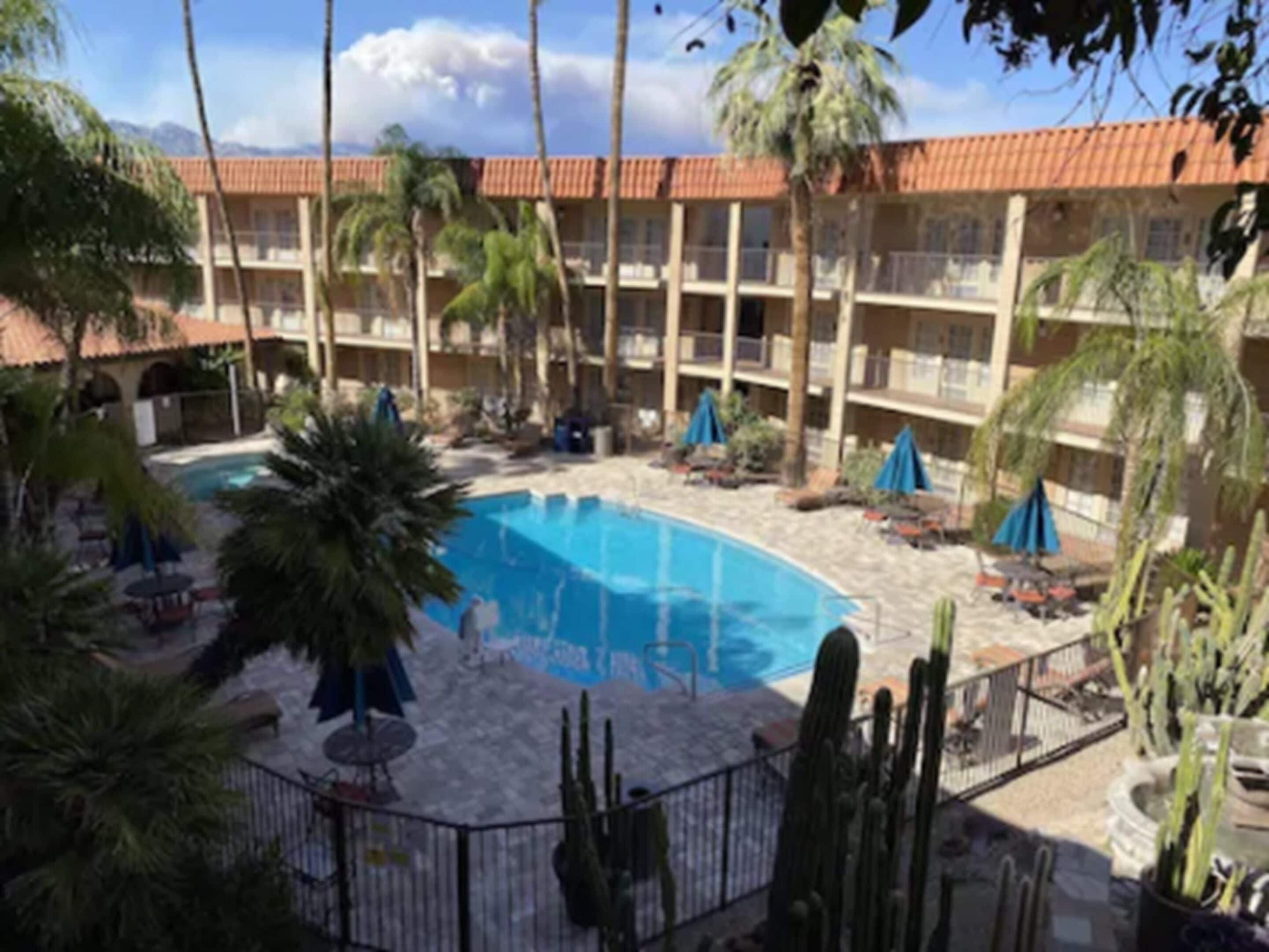 A hotel with a central swimming pool surrounded by palm trees and cacti, with multiple balconies and a red-tiled roof.