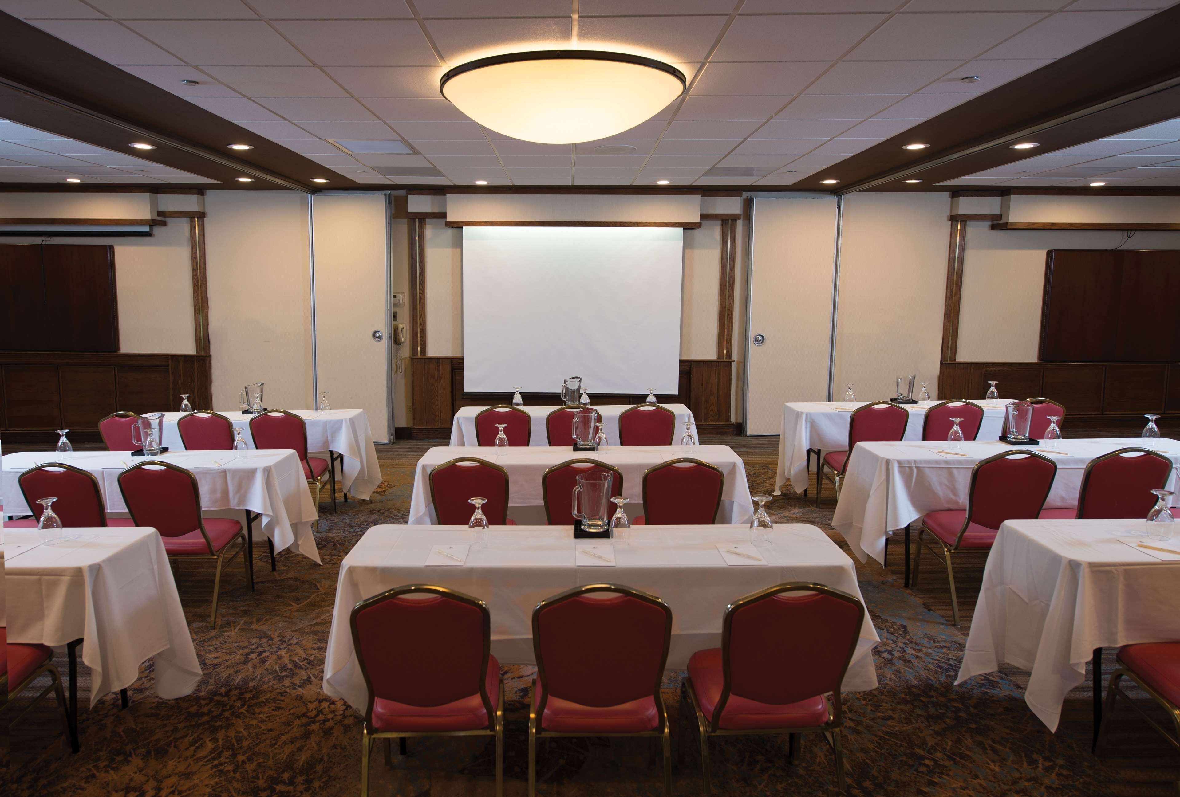 A conference room with multiple tables and red chairs, a projector screen, and a large chandelier.