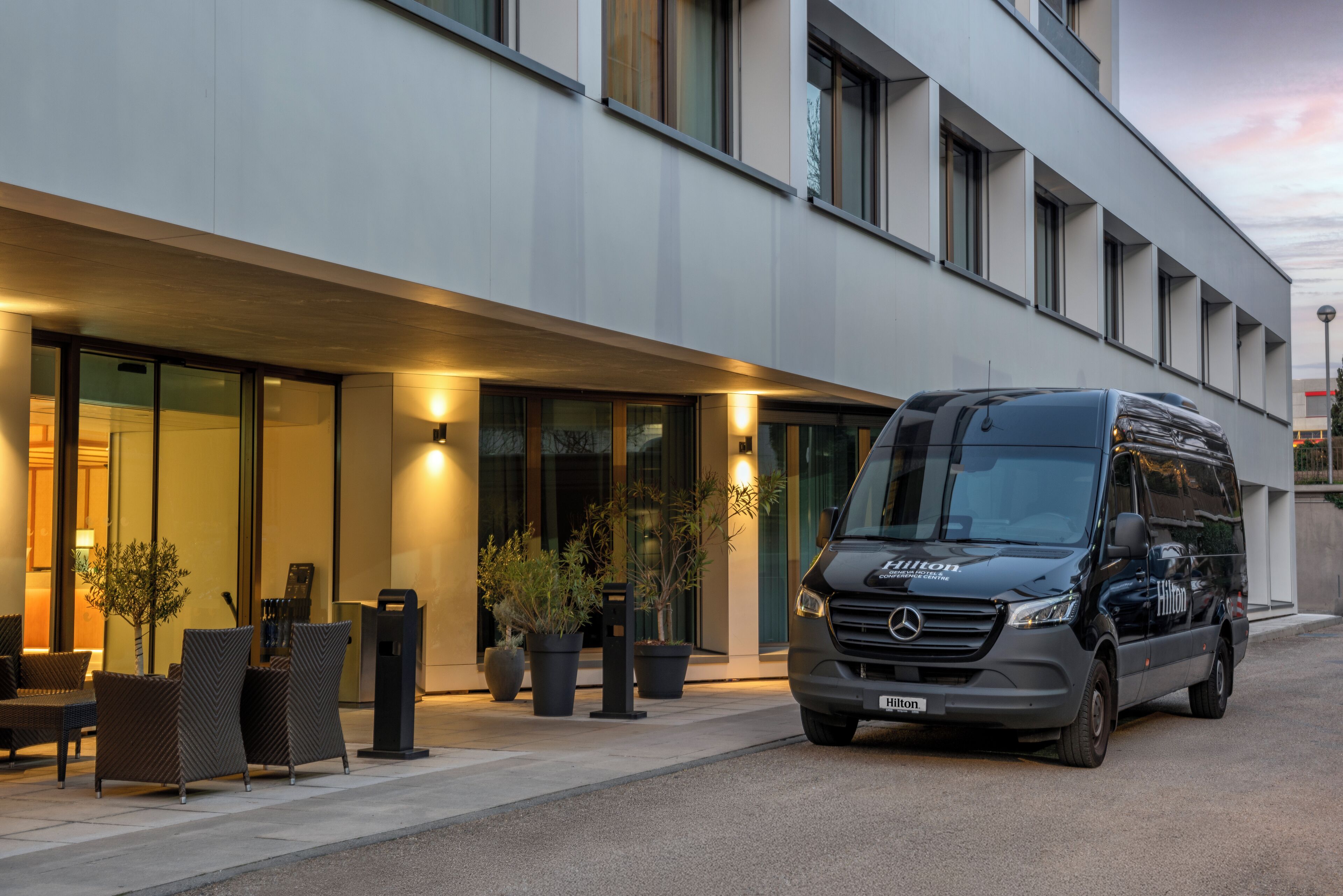 A modern building with a Mercedes-Benz van parked in front, large windows, and outdoor seating.