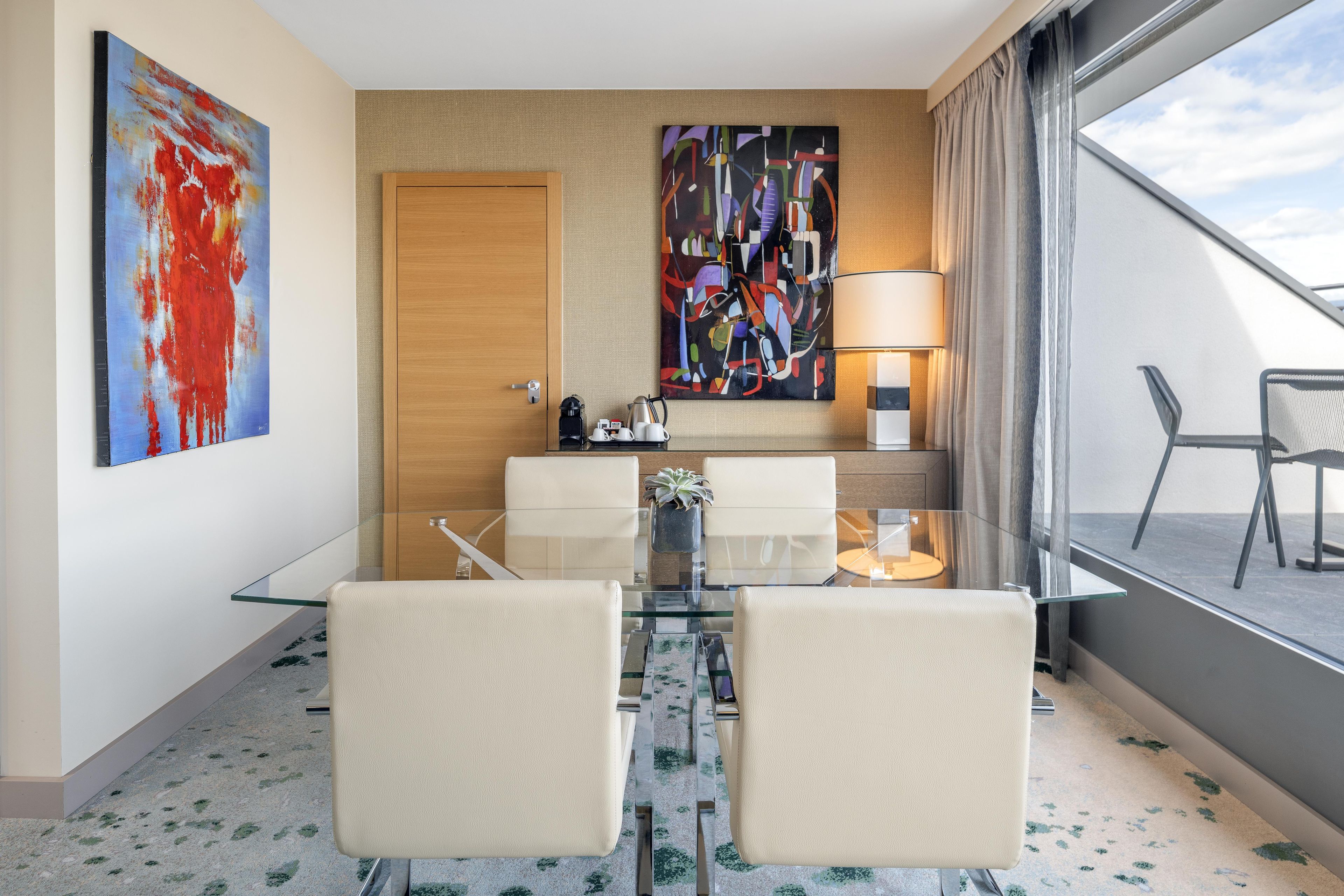 Quarto de hotel com mesa de vidro, duas cadeiras, sofá, máquina de café, chaleira, planta e obras de arte nas paredes.