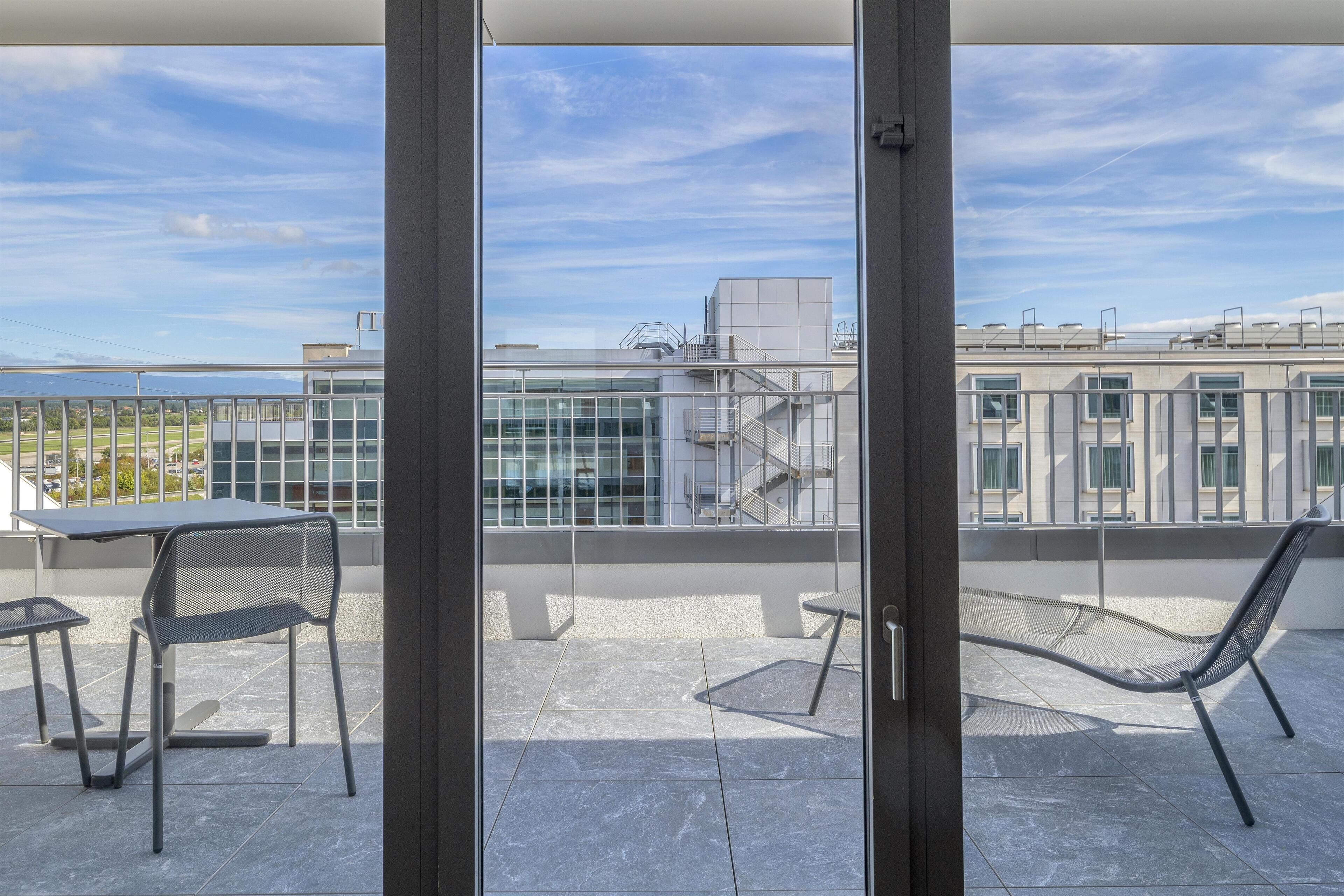 A balcony with a table and chairs, a hammock, and a view of a building and sky.