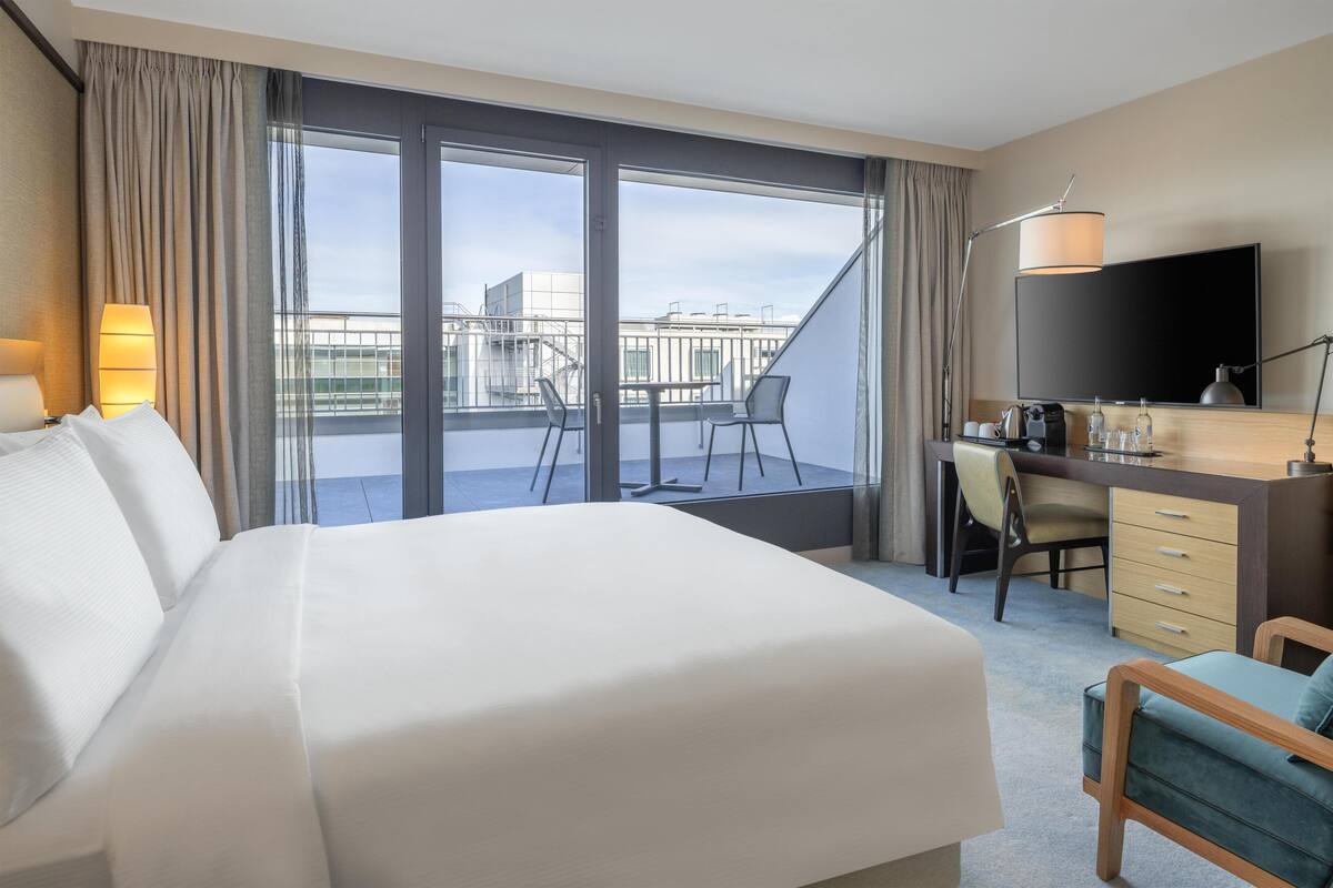 A hotel room with a large bed, a desk with a chair, a television, and a balcony with a view of the city.