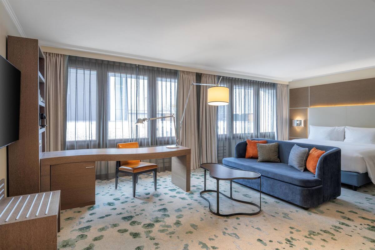 A hotel room with a desk, a sofa, a bed, a TV, and large windows with curtains.