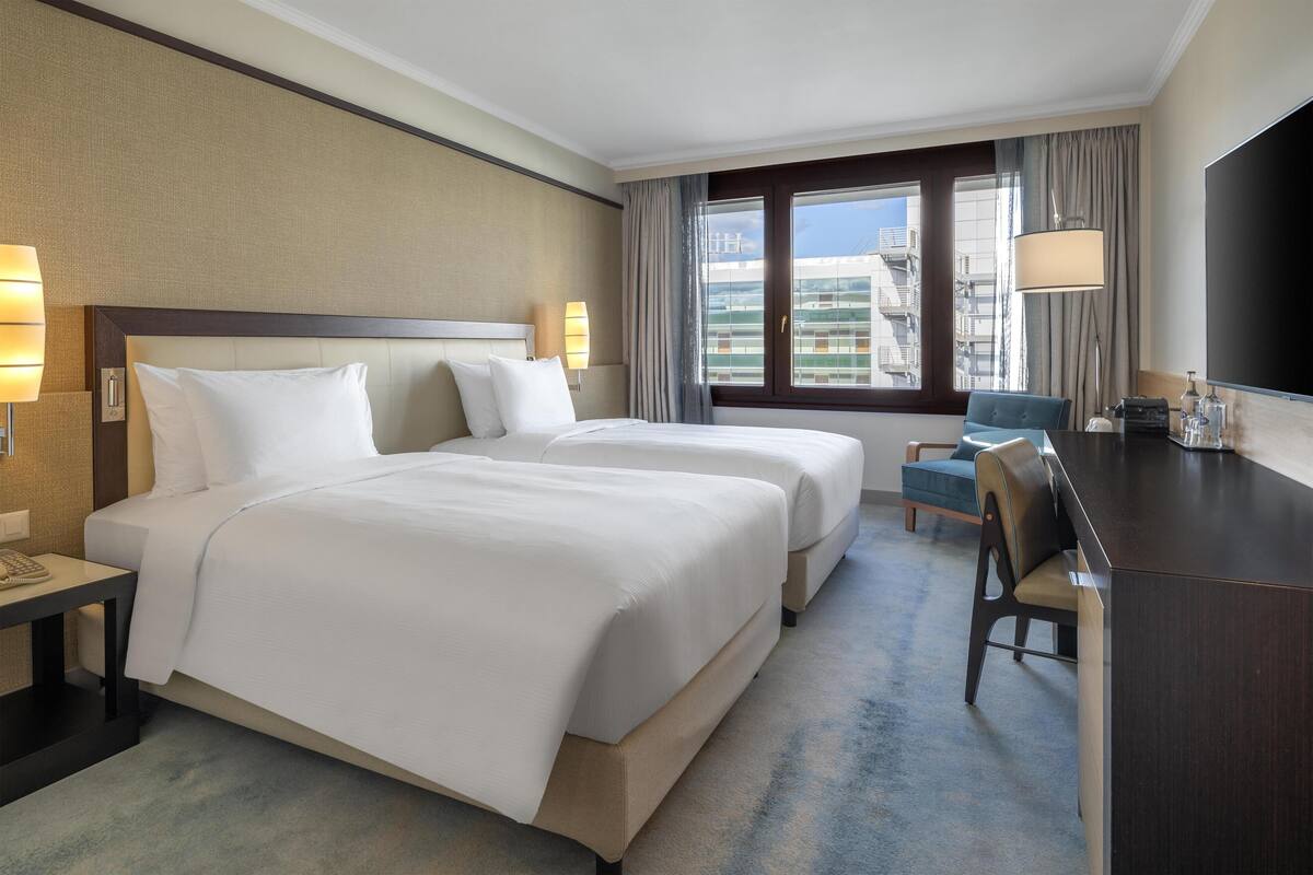 A hotel room with two beds, a desk, a chair, a TV, and a window with a city view.