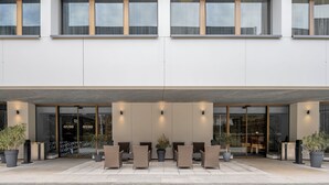 A modern building with a covered entrance, large windows, and outdoor seating area with potted plants. - Hilton Geneva Hotel and Conference Centre (Le Grand-Saconnex)