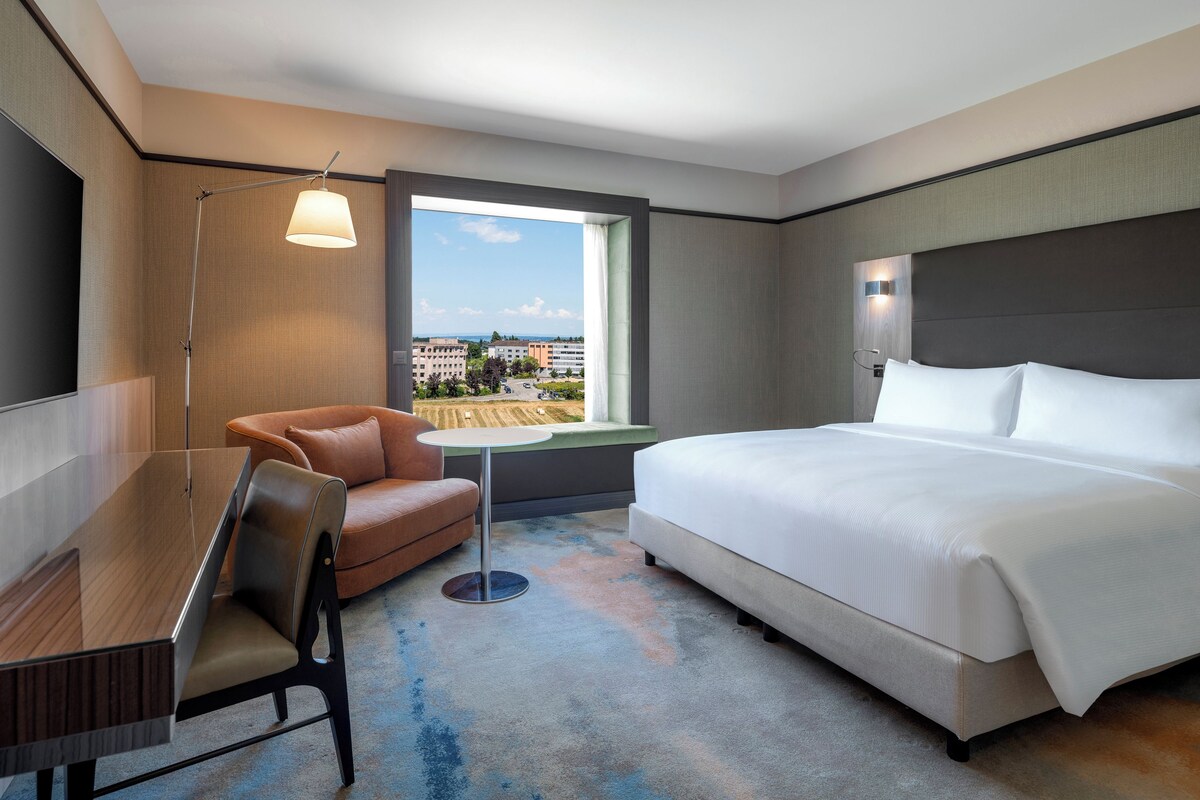 A hotel room with a bed, a desk, a chair, a small table, and a view of the cityscape.