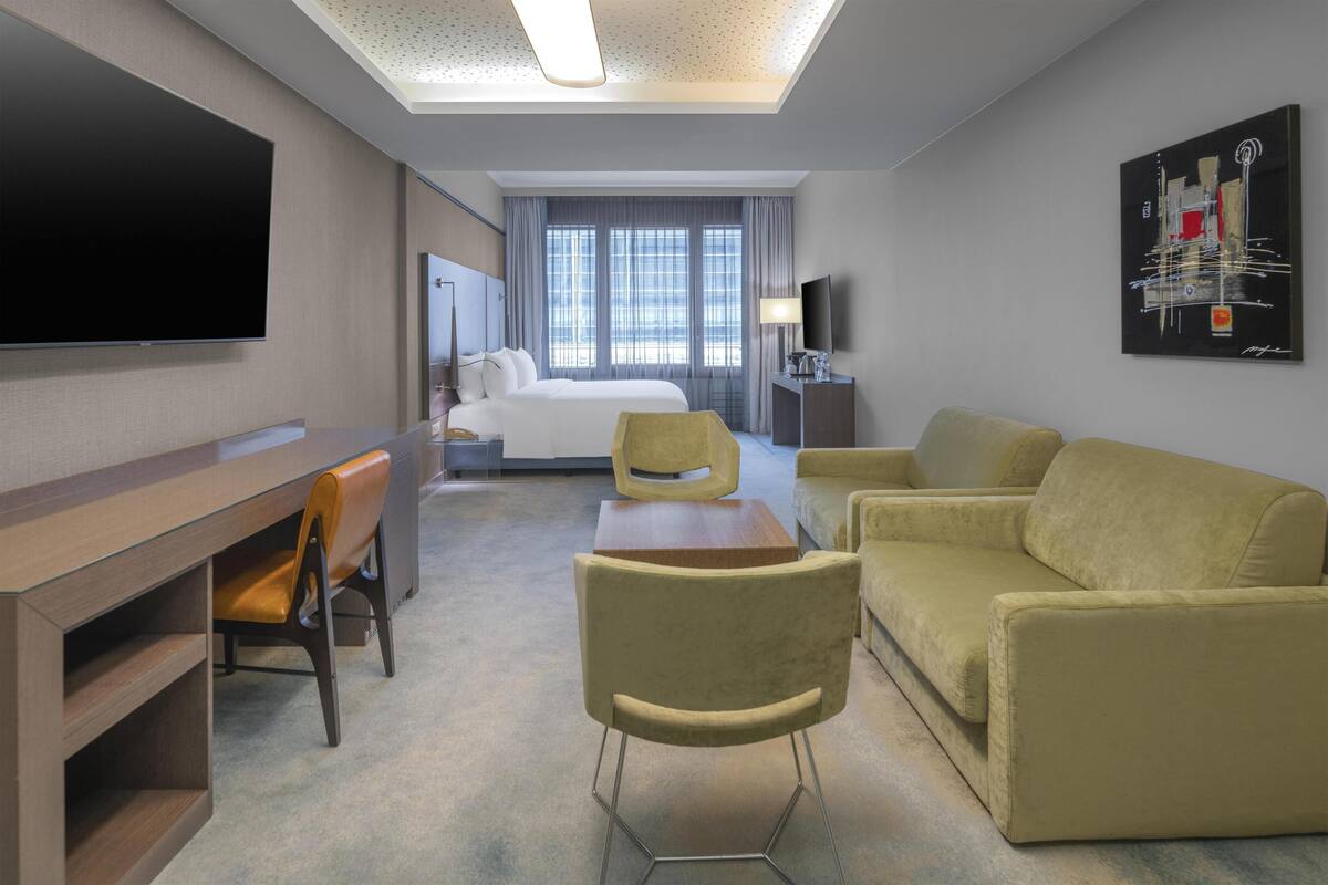 A hotel room with a bed, a desk with a chair, a sofa, a TV, and a painting on the wall.