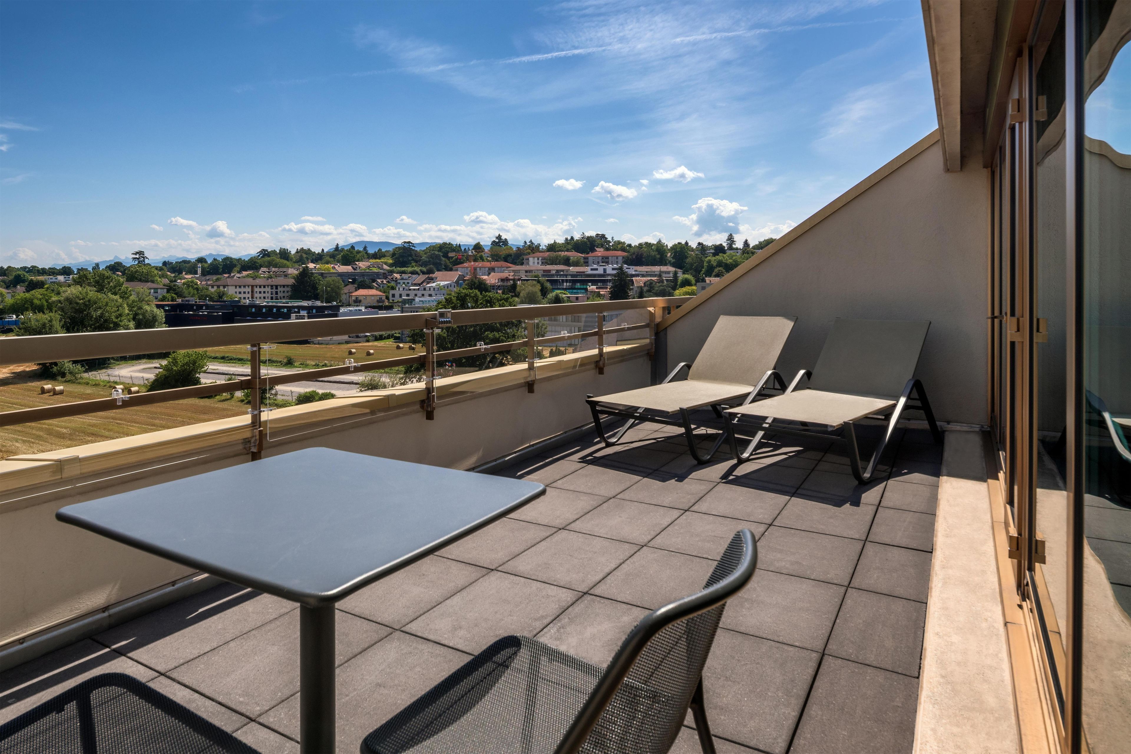 A balcony with a table and two chairs, overlooking a residential area with houses and a road.